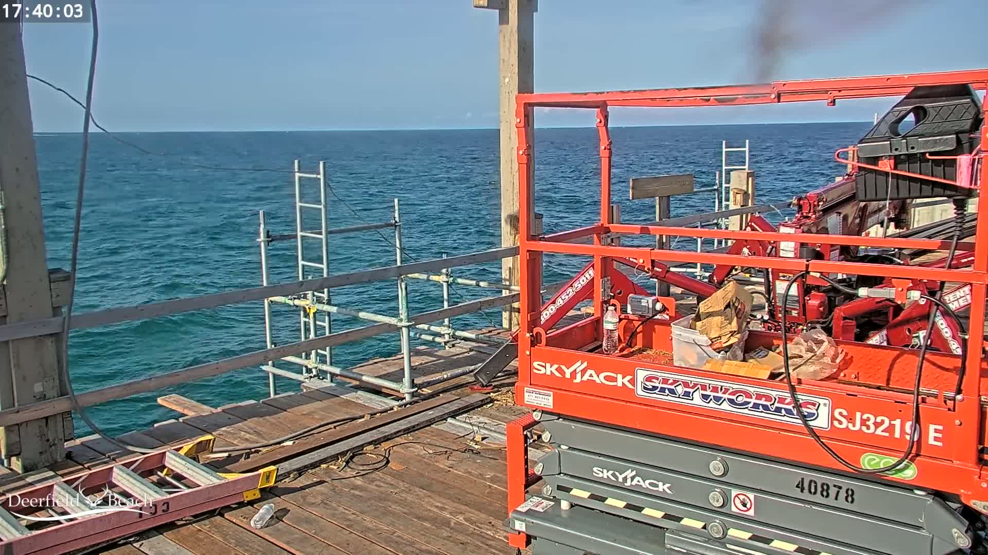 A red scissor lift sits on a wooden pier overlooking a calm blue ocean under a clear sky.