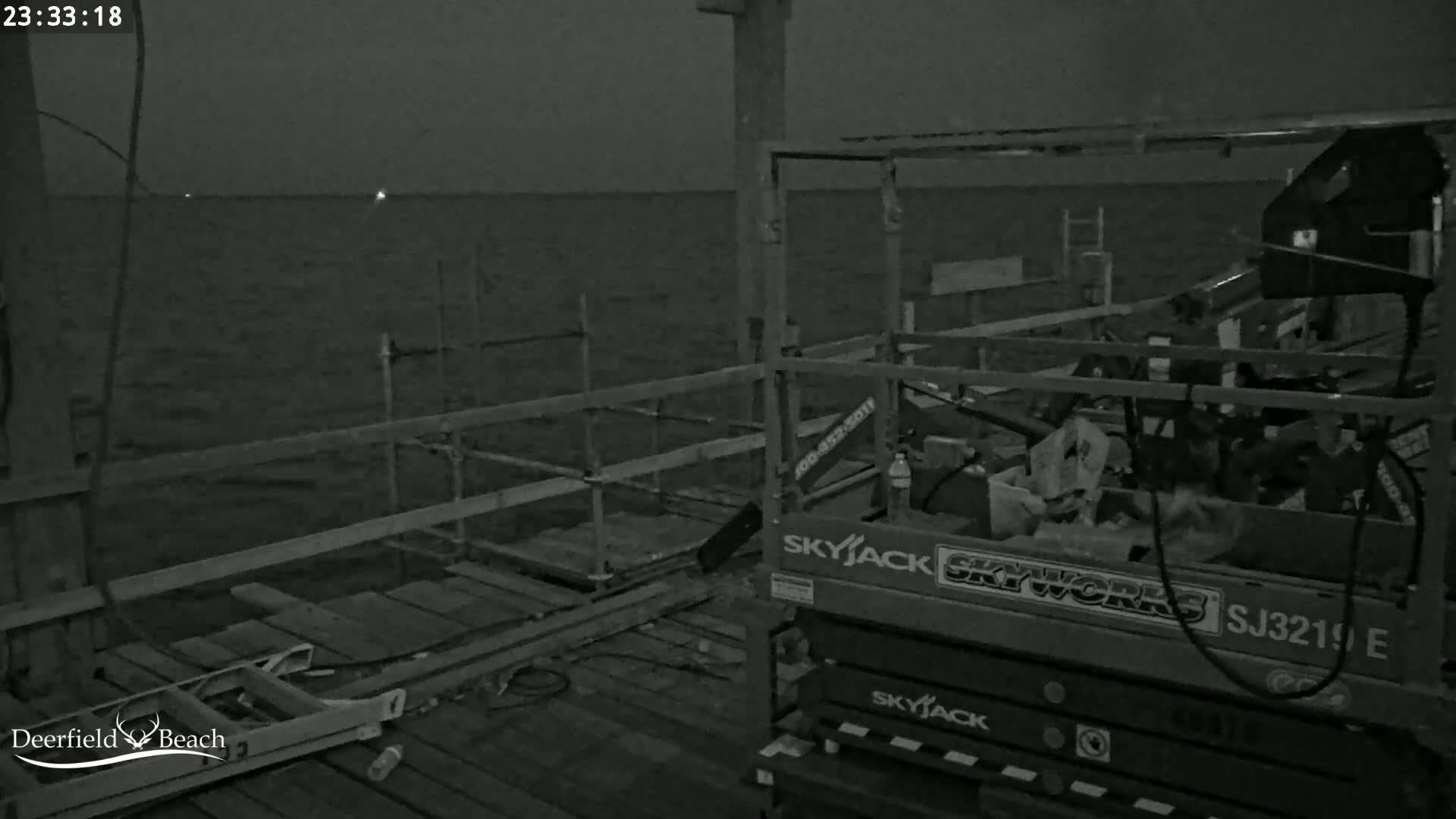 A dark nighttime scene shows a section of a pier under construction with scaffolding, a lift, and a calm body of water extending to a dark horizon.
