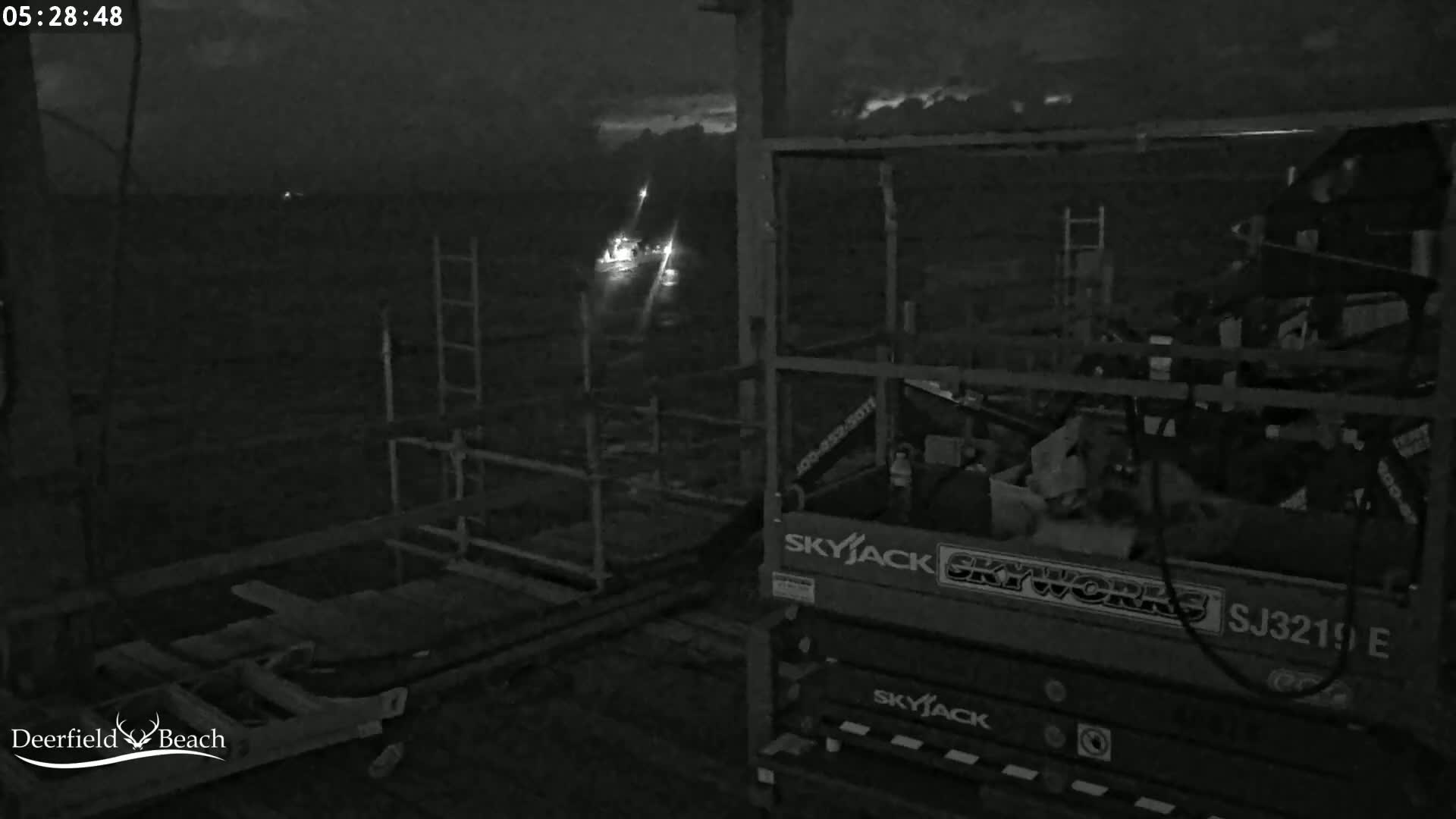 A nighttime, low-light image shows a construction or maintenance site with a scissor lift and some scaffolding, with distant lights visible in the background under a dark, cloudy sky.