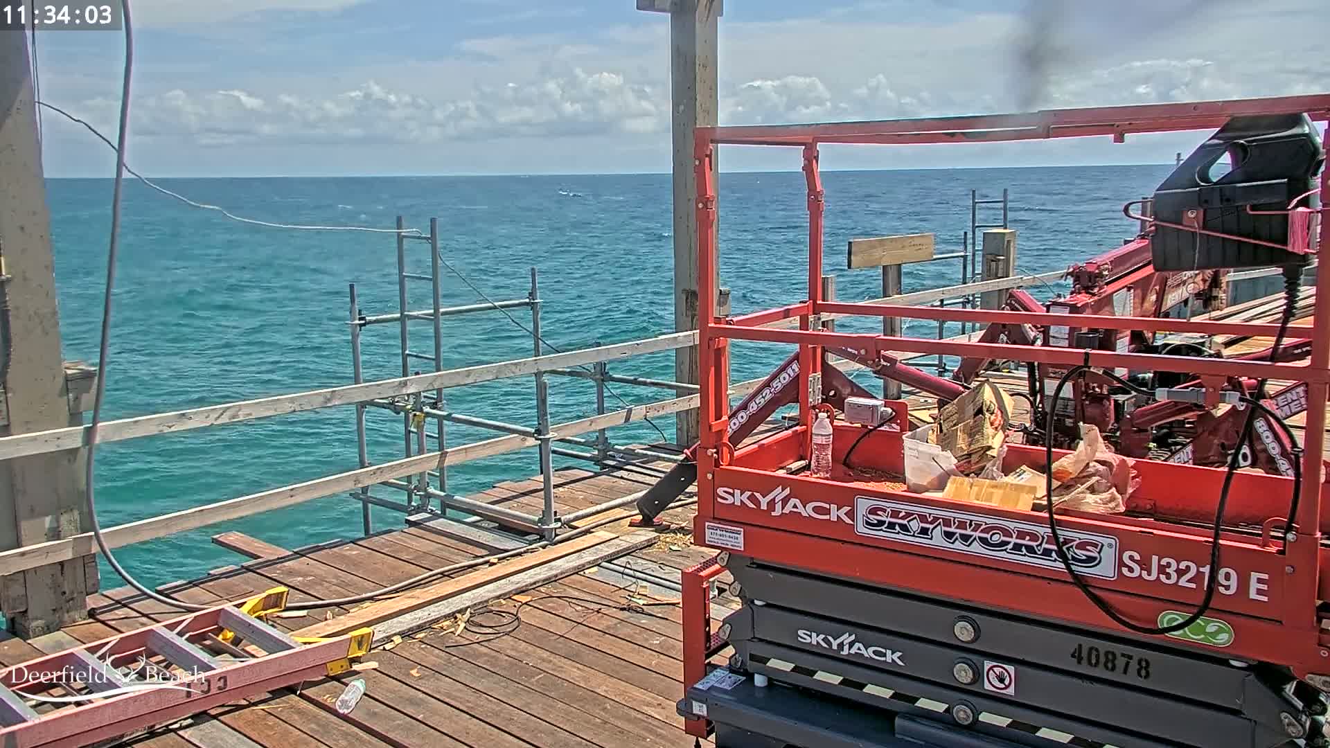 A red Skyjack lift is parked on a wooden pier overlooking a blue ocean under a partly cloudy sky.