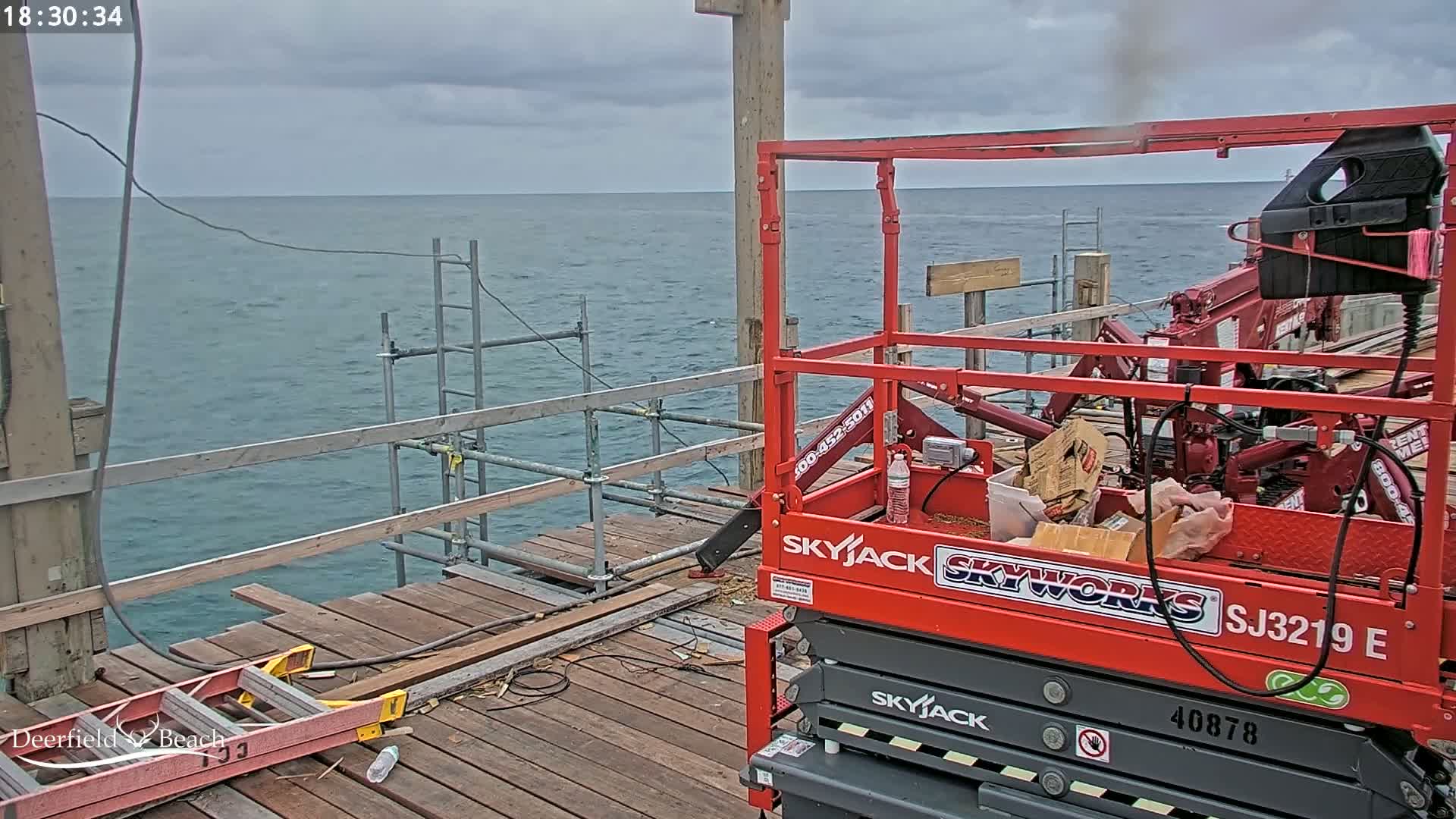 A red scissor lift sits on a weathered wooden pier overlooking a calm, gray ocean under an overcast sky.