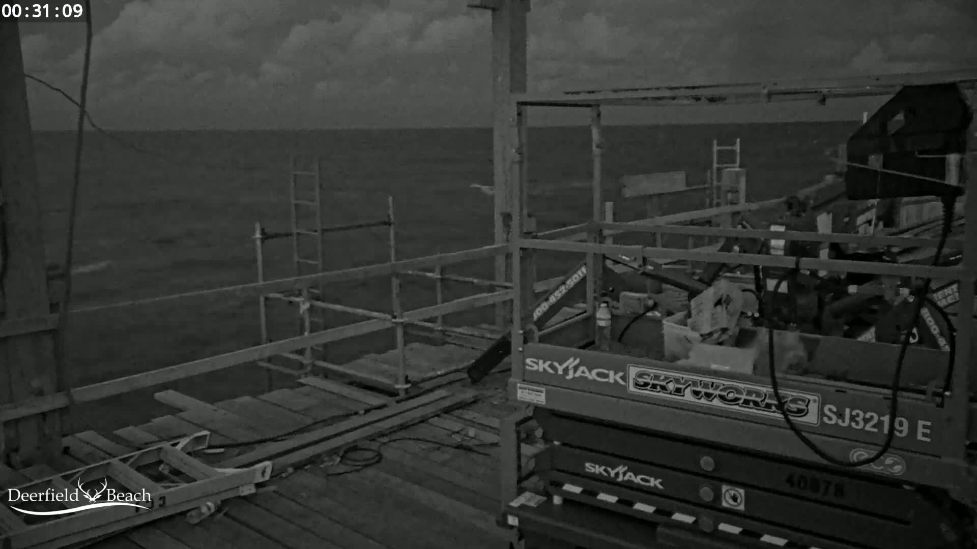 A dark, overcast night scene shows a section of a pier under construction, with a scissor lift and scaffolding visible, overlooking a choppy sea.
