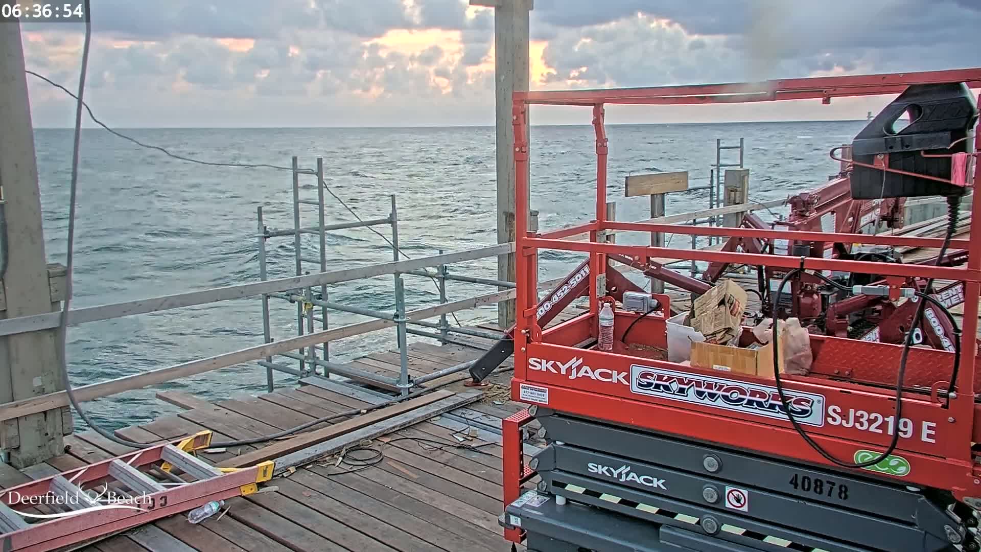 A red Skyjack lift is on a wooden pier overlooking a choppy sea under a cloudy sky.
