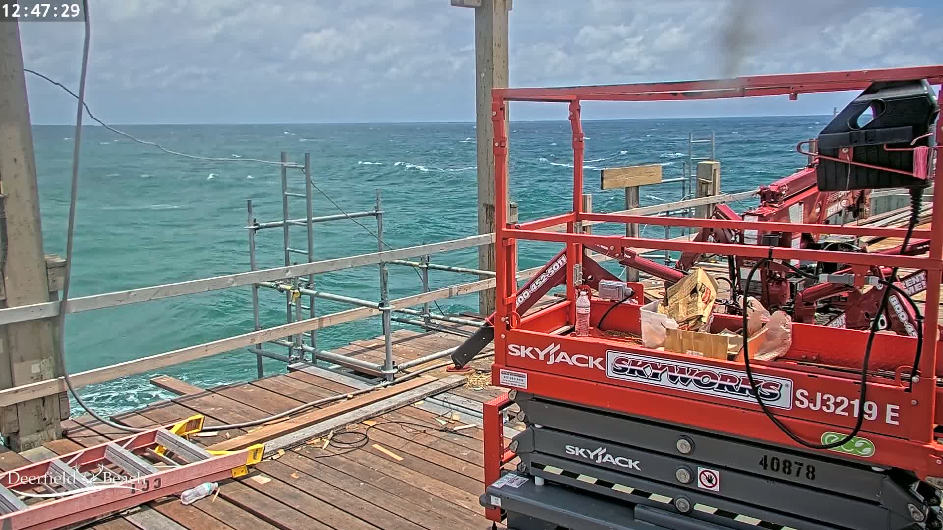 A red Skyjack lift is parked on a wooden pier overlooking a choppy teal ocean under a partly cloudy sky.