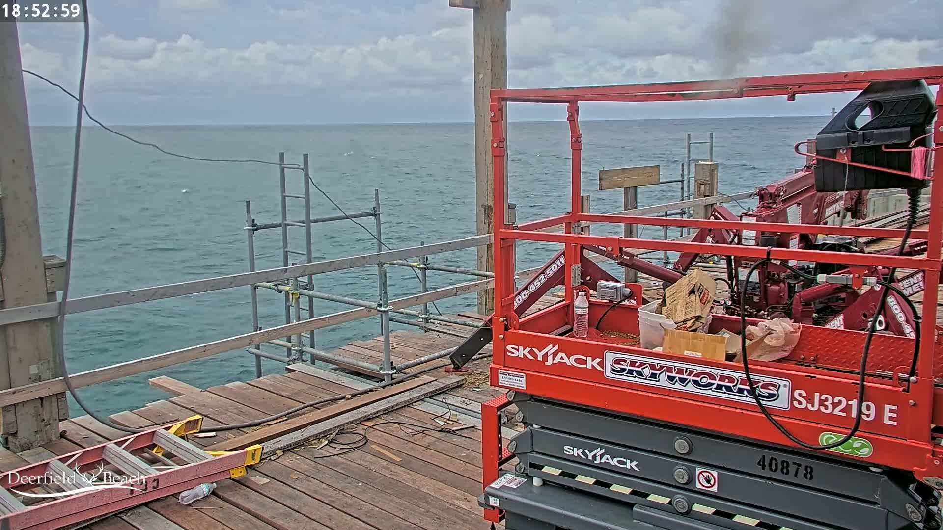 A red scissor lift sits on a wooden pier overlooking a calm, gray-blue ocean under a partly cloudy sky.