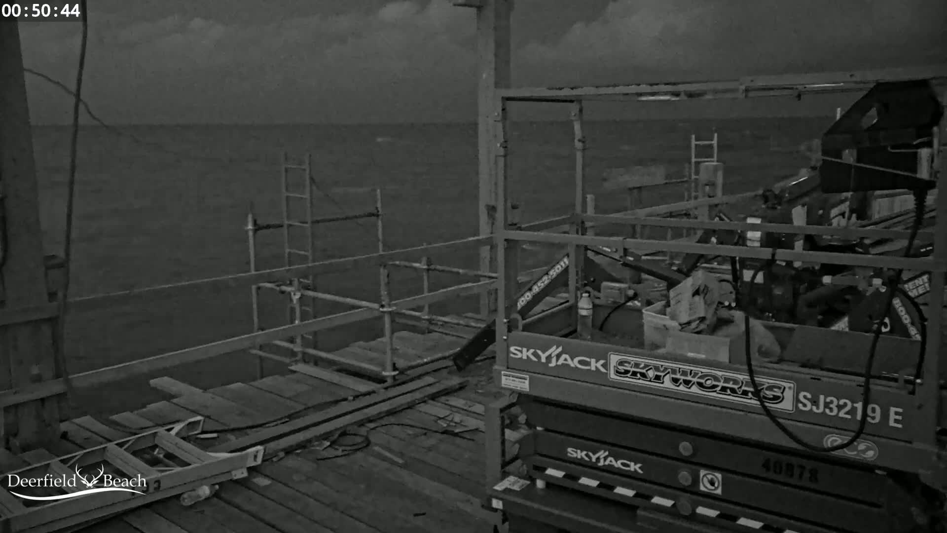 A nighttime, overcast scene shows a body of water, a wooden pier with scaffolding and a scissor lift.