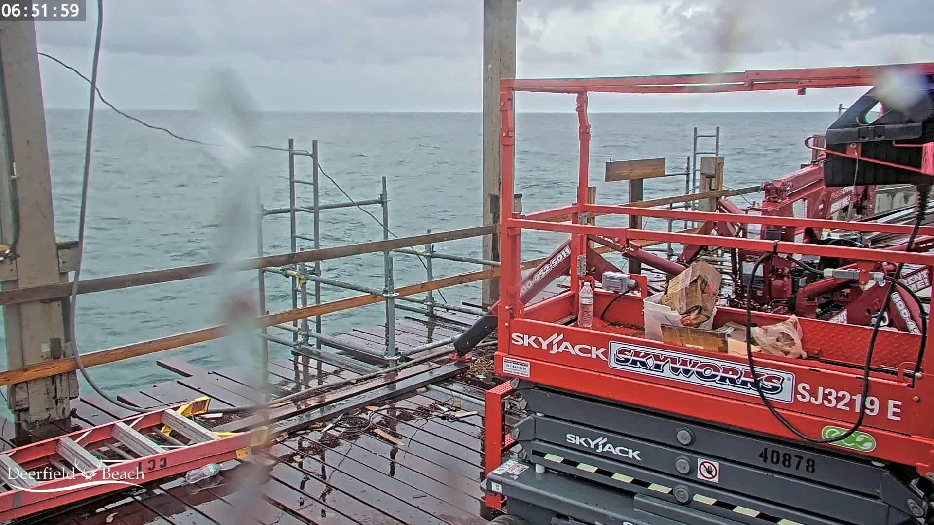 A red Skyjack lift is parked on a rain-soaked pier overlooking a choppy ocean under a cloudy sky.
