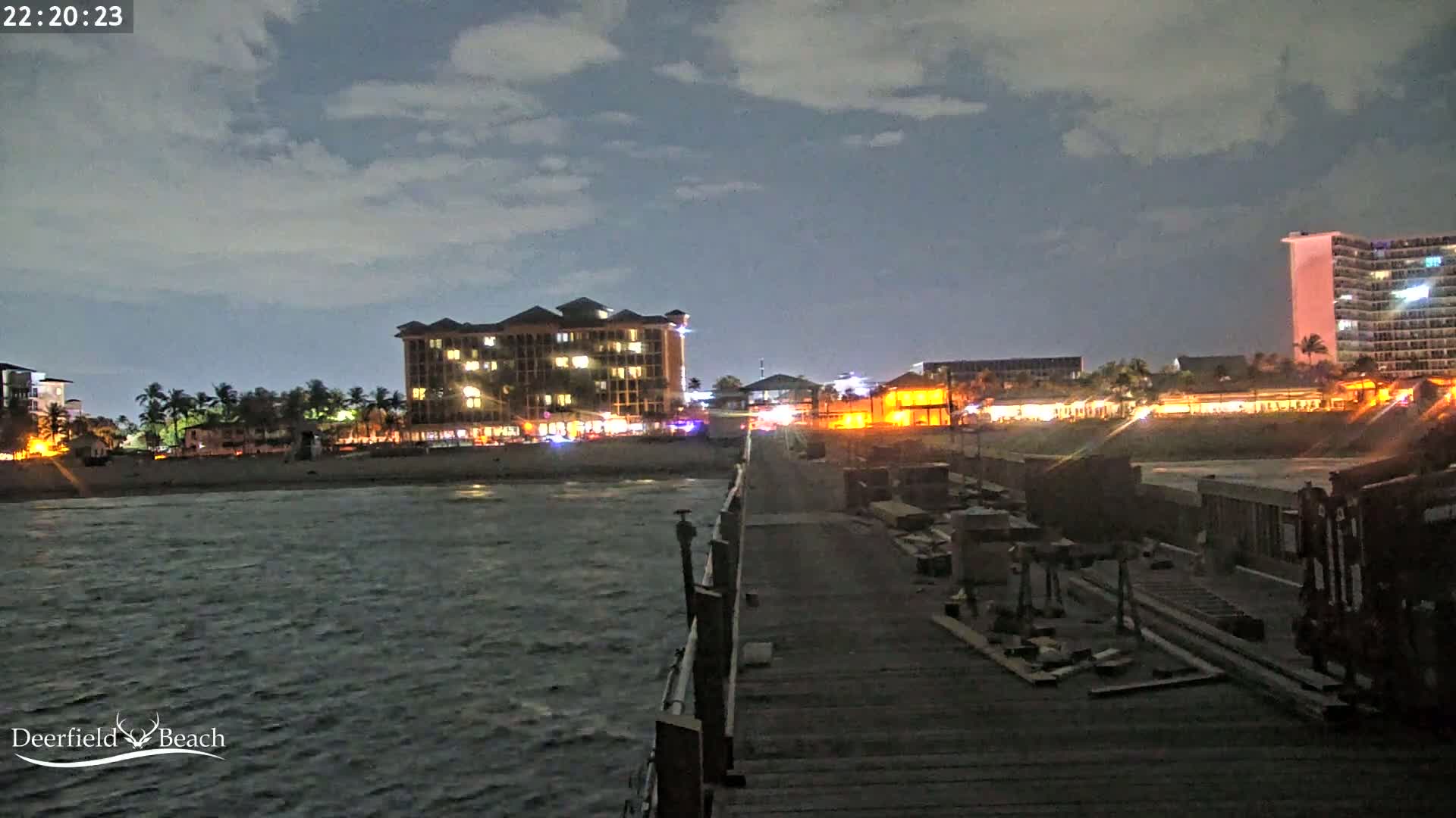 A nighttime view of a beach with illuminated buildings, palm trees, and a pier under a partly cloudy sky.