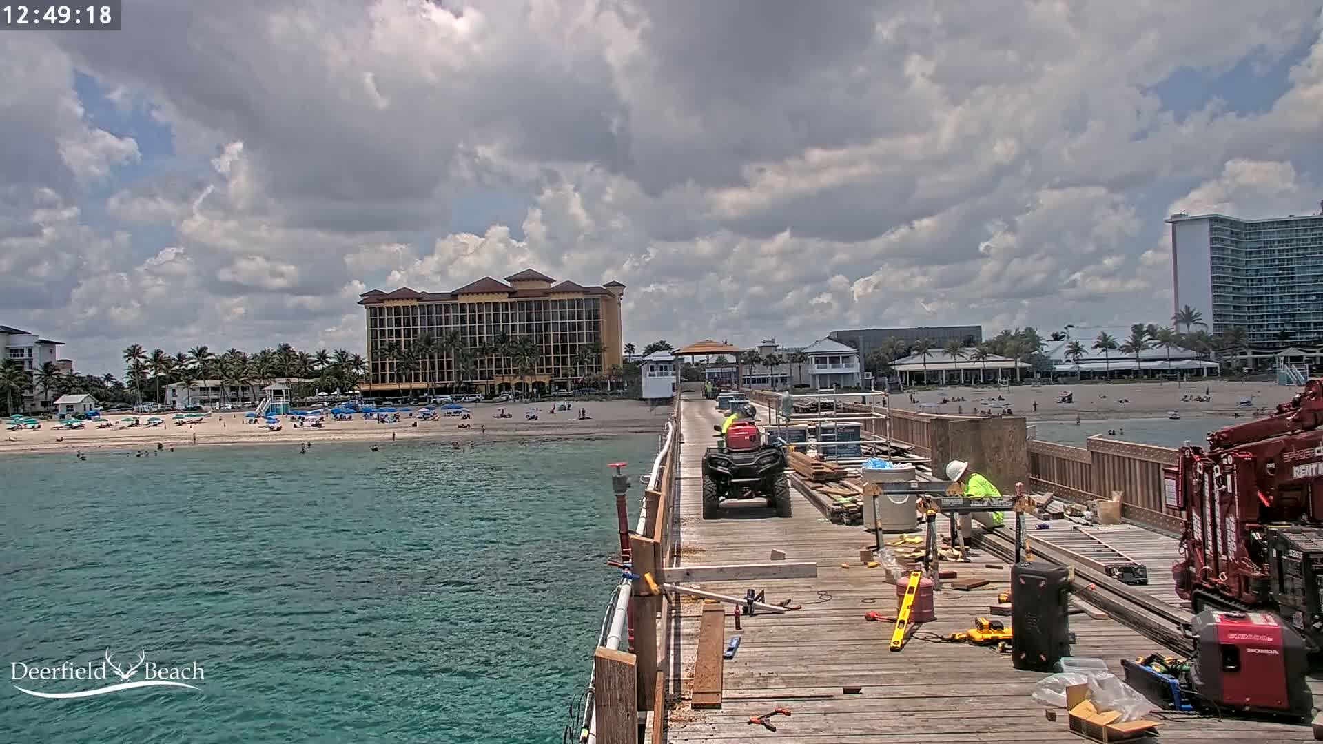 Construction workers are repairing a pier overlooking a beach with turquoise water under a partly cloudy sky.