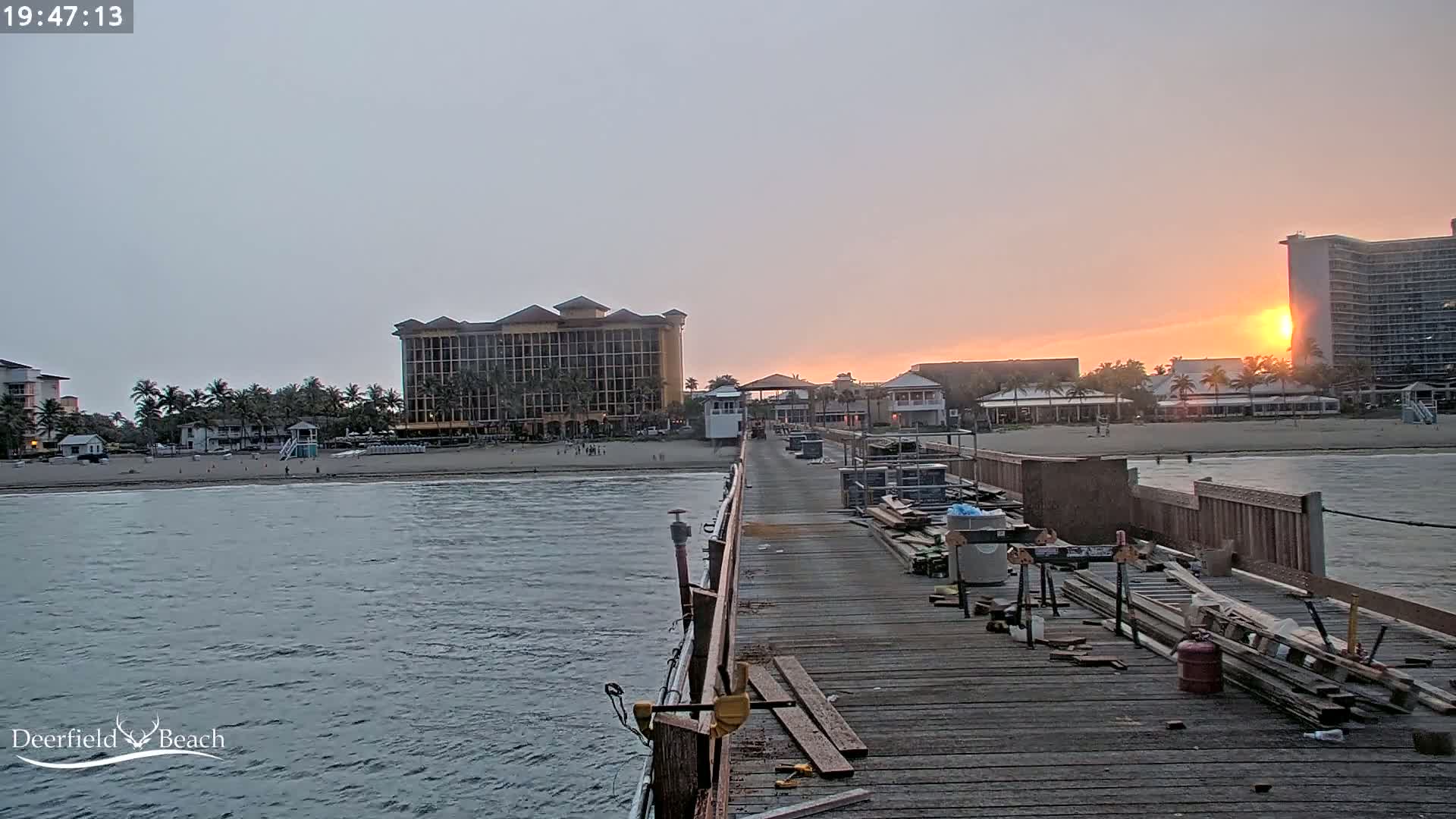 A long wooden pier extends from the viewer's perspective toward a beach, hotels, and the setting sun during a calm evening.
