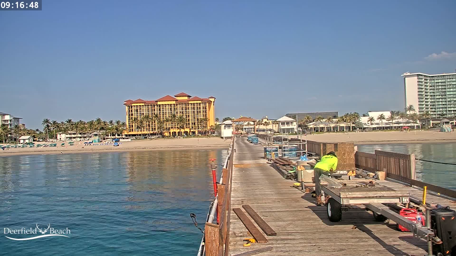 A worker on a pier near a beach with calm water, under a clear blue sky, is working on what appears to be some kind of construction or repair.