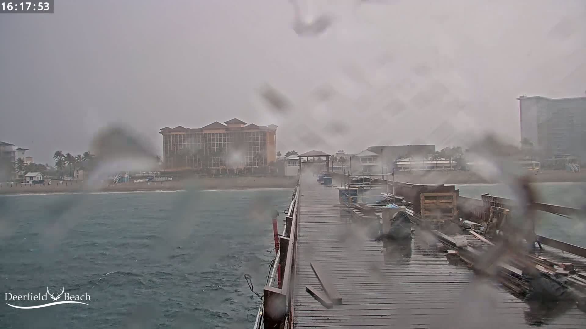 A rain-soaked pier extends into a choppy ocean toward a hotel and other buildings during a heavy rainstorm.
