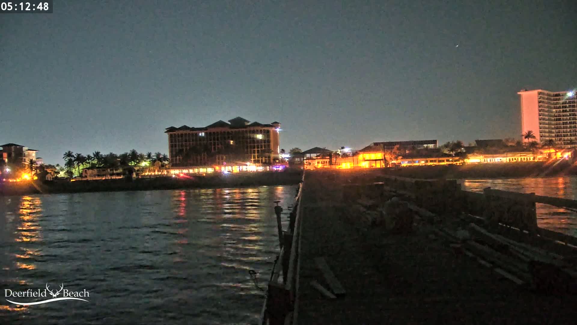 A nighttime view from a pier shows illuminated buildings along a waterfront, with calm water reflecting the lights.