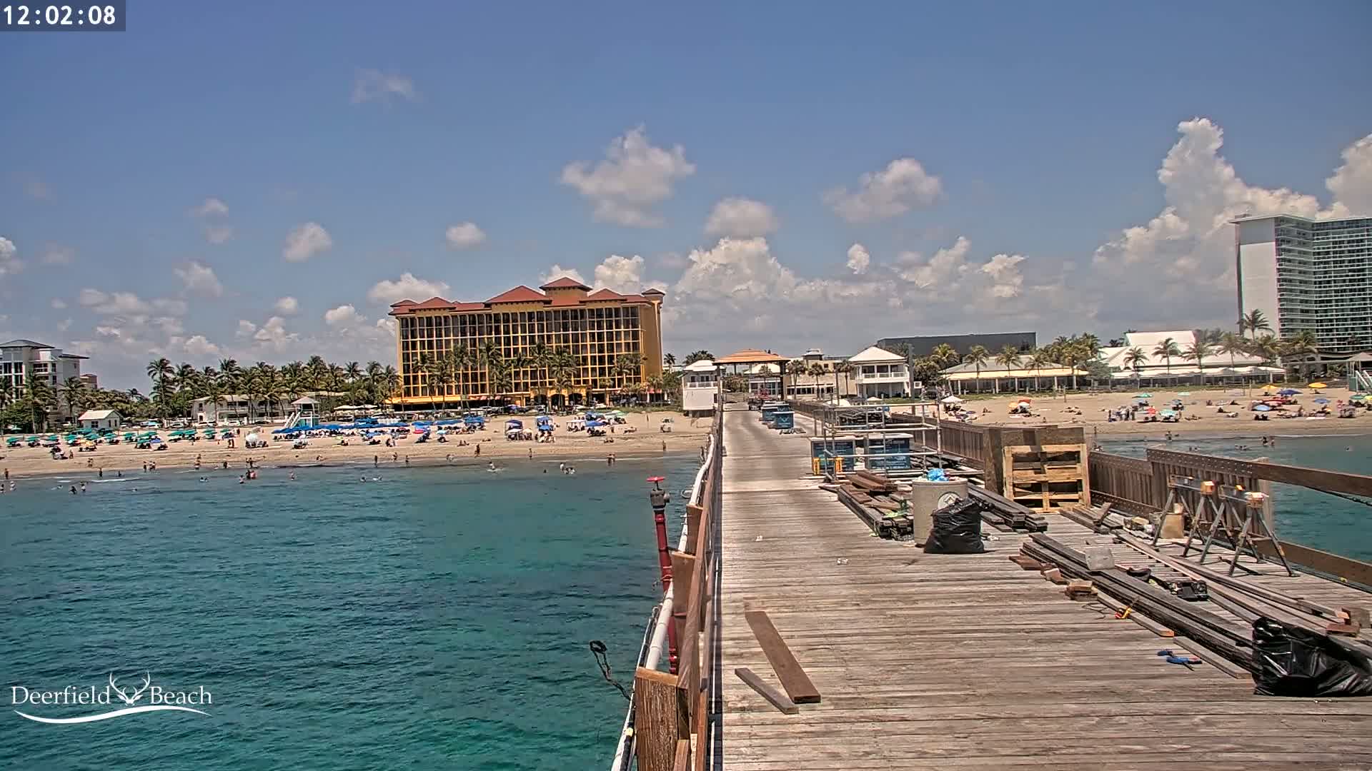 A wooden pier extends over teal water toward a sandy beach with people, beach umbrellas, and buildings under a partly cloudy sky.