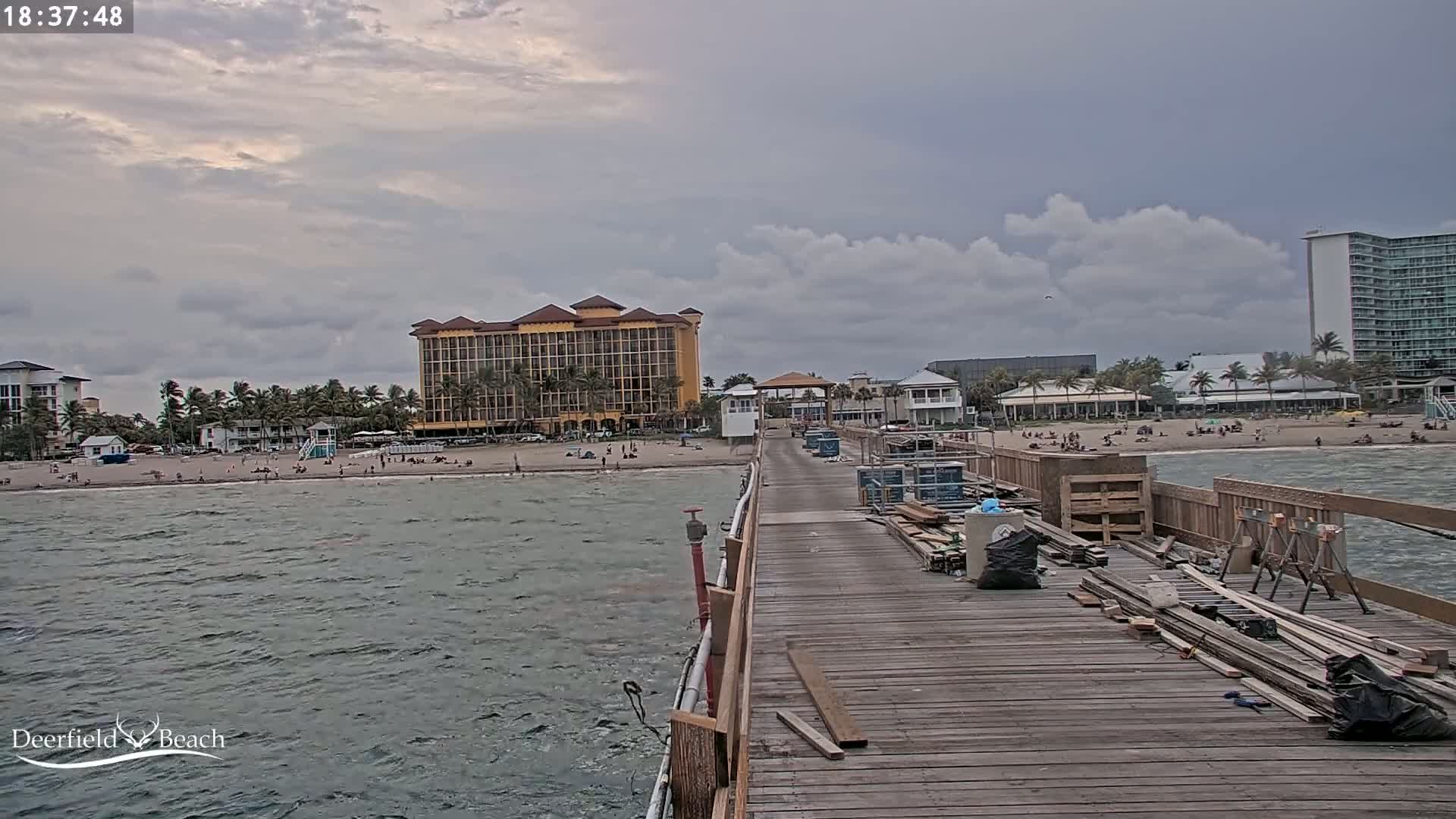 A long wooden pier extends over a choppy ocean towards a beach with people and buildings under a cloudy sky.