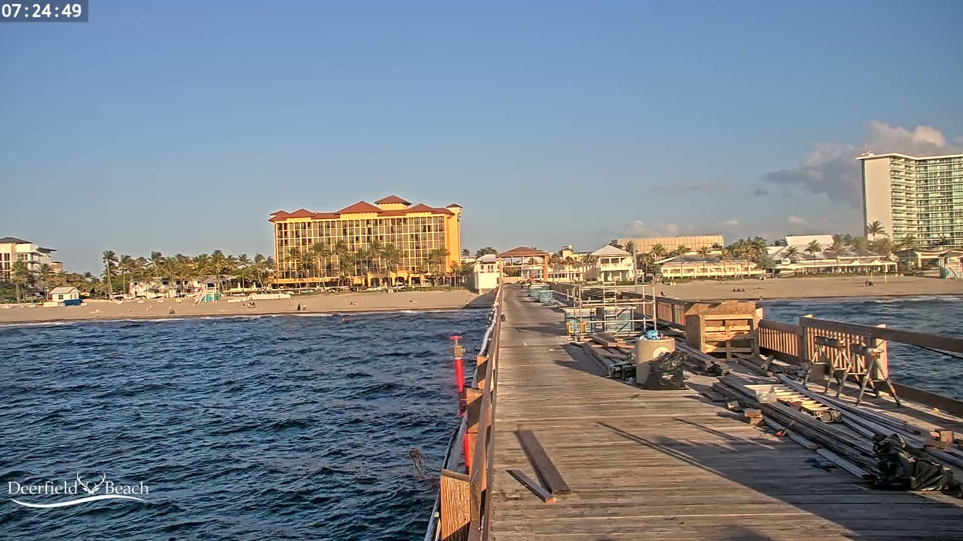 A wooden pier extends over a wavy ocean towards a sandy beach with buildings and palm trees under a mostly clear sky.