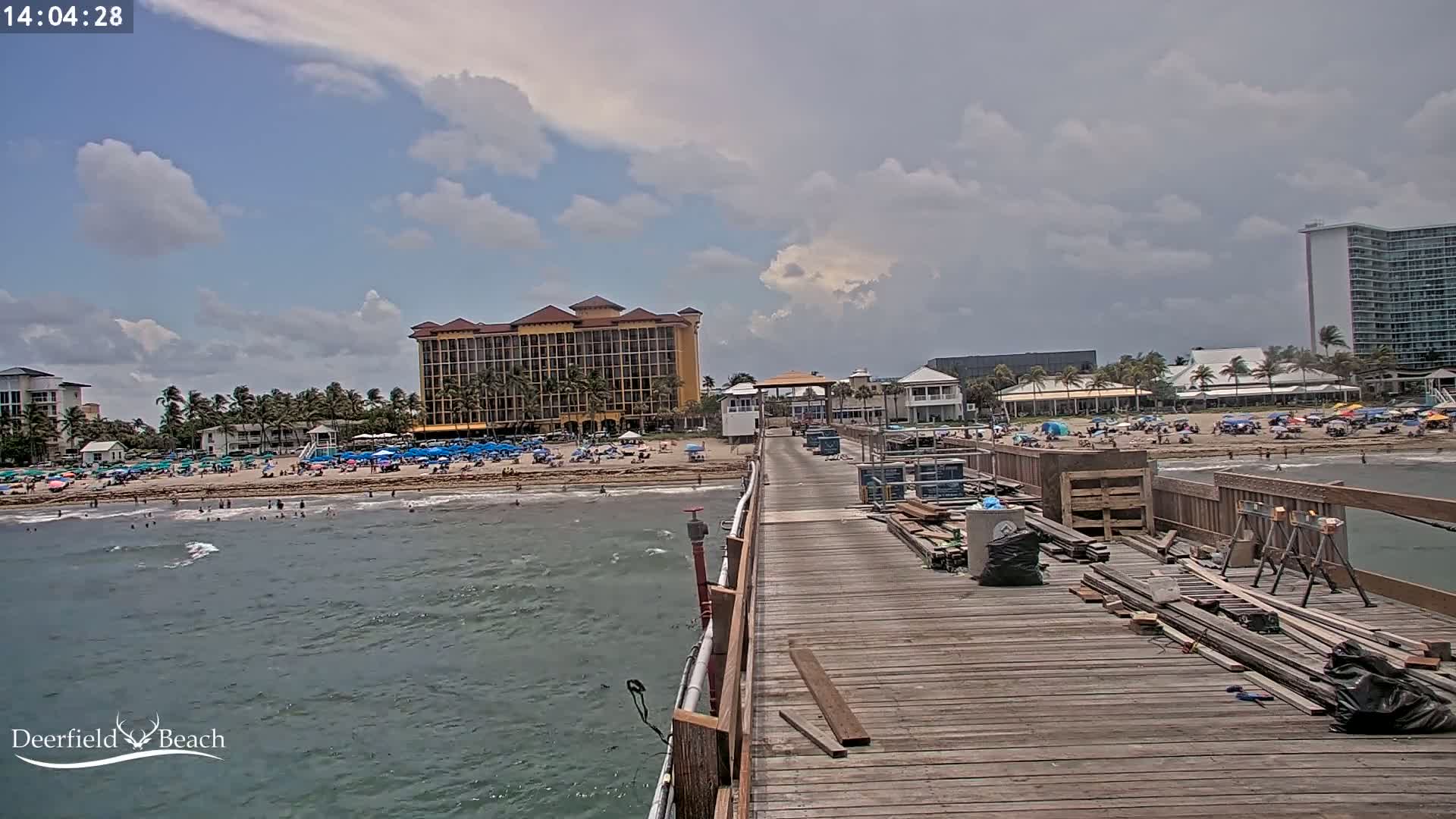A partly cloudy day at a beach with a crowded shoreline, a pier under construction, and several high-rise buildings in the background.