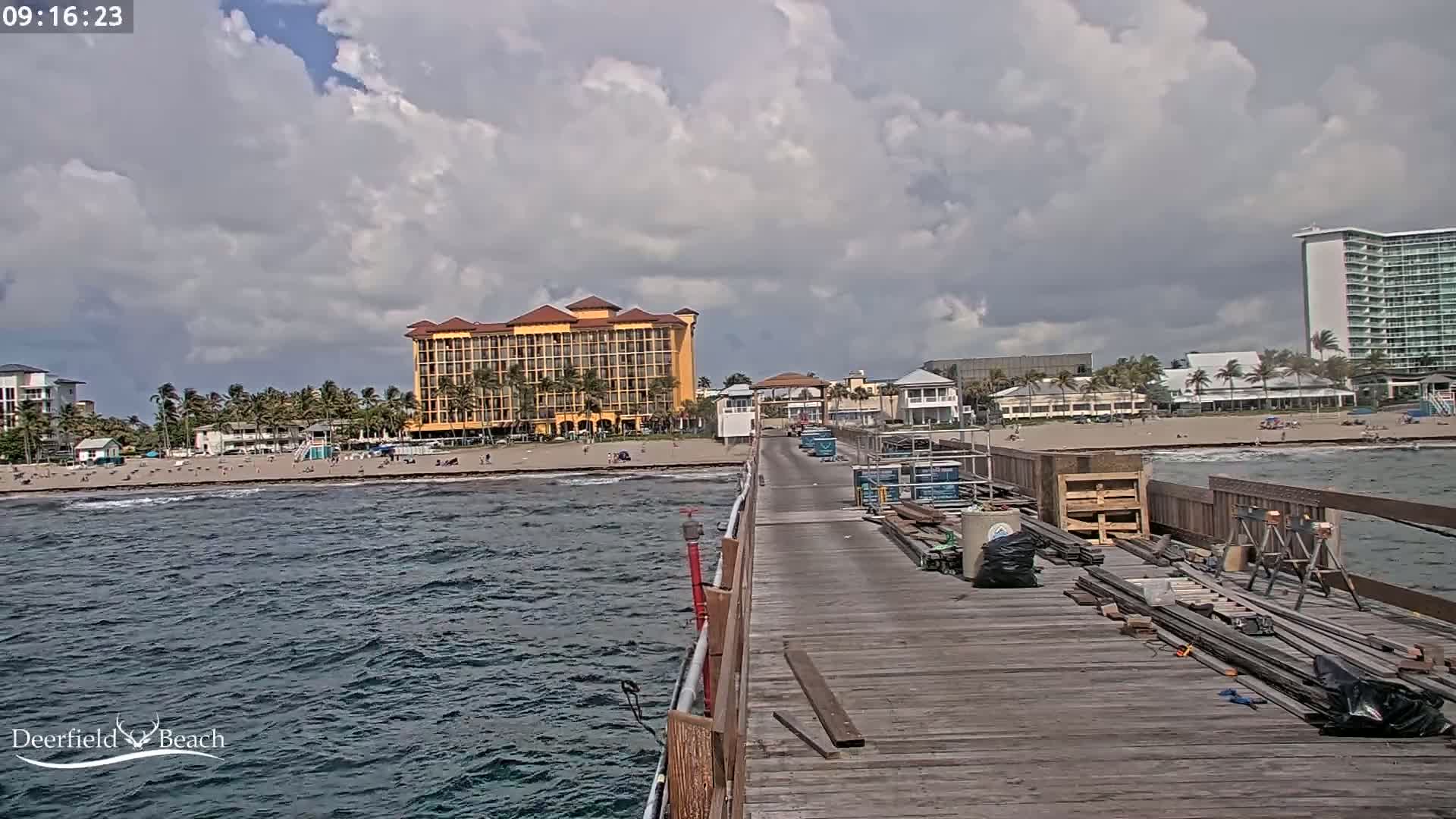 A partly cloudy day shows a view from a pier over a beach with several buildings, including a large yellow hotel and a tall white high-rise, with construction materials on the pier.