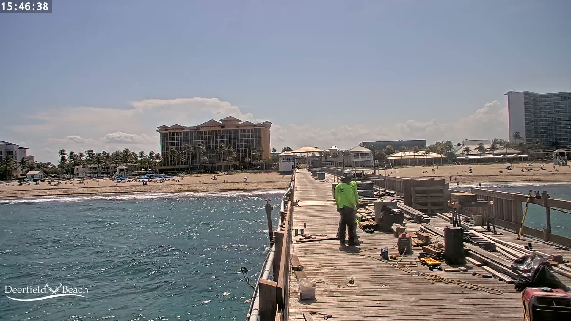 A worker stands on a pier under a partly cloudy sky, overlooking a beach with people and several buildings in the background.