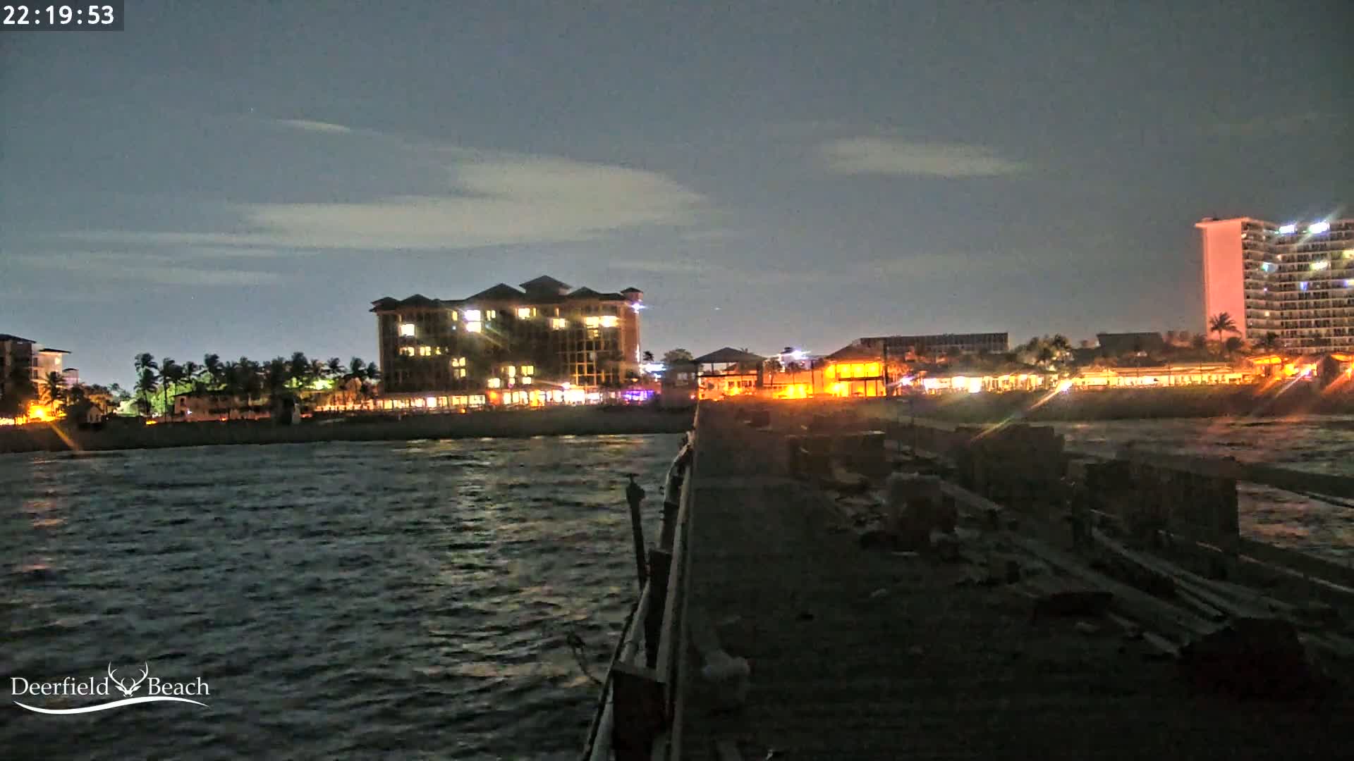 A nighttime view from a pier shows a dark, wavy ocean and a shoreline with illuminated buildings under a partly cloudy sky.