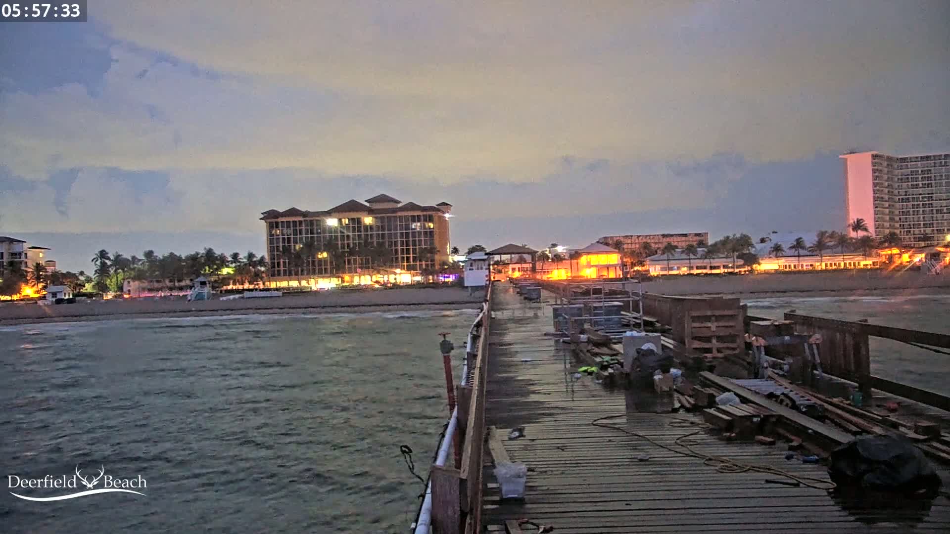 A long pier extends from a beach at dusk, with buildings and palm trees lining the shore under a mostly cloudy sky.