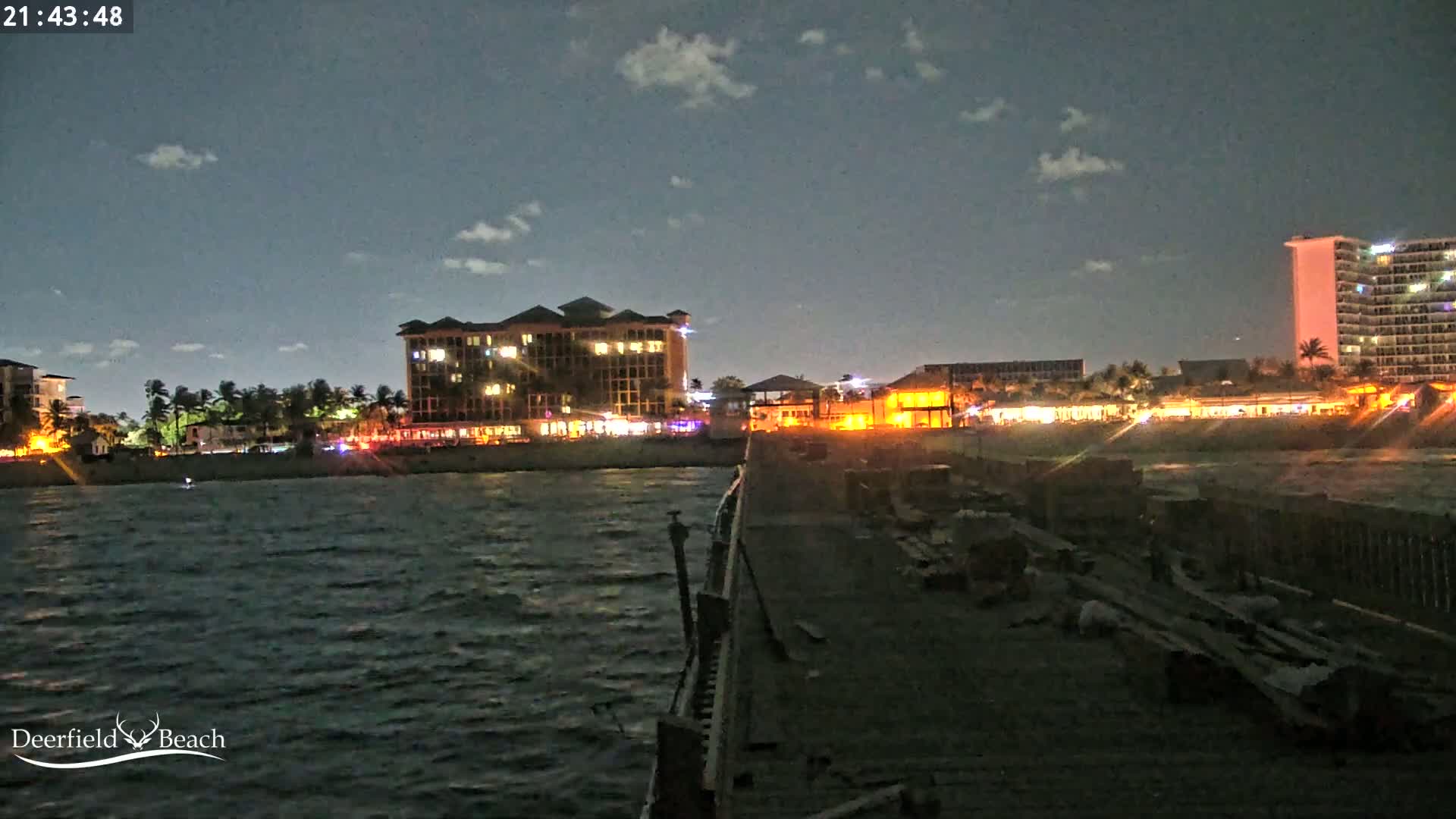 A nighttime view from a pier shows a shoreline with illuminated buildings and a slightly choppy ocean.