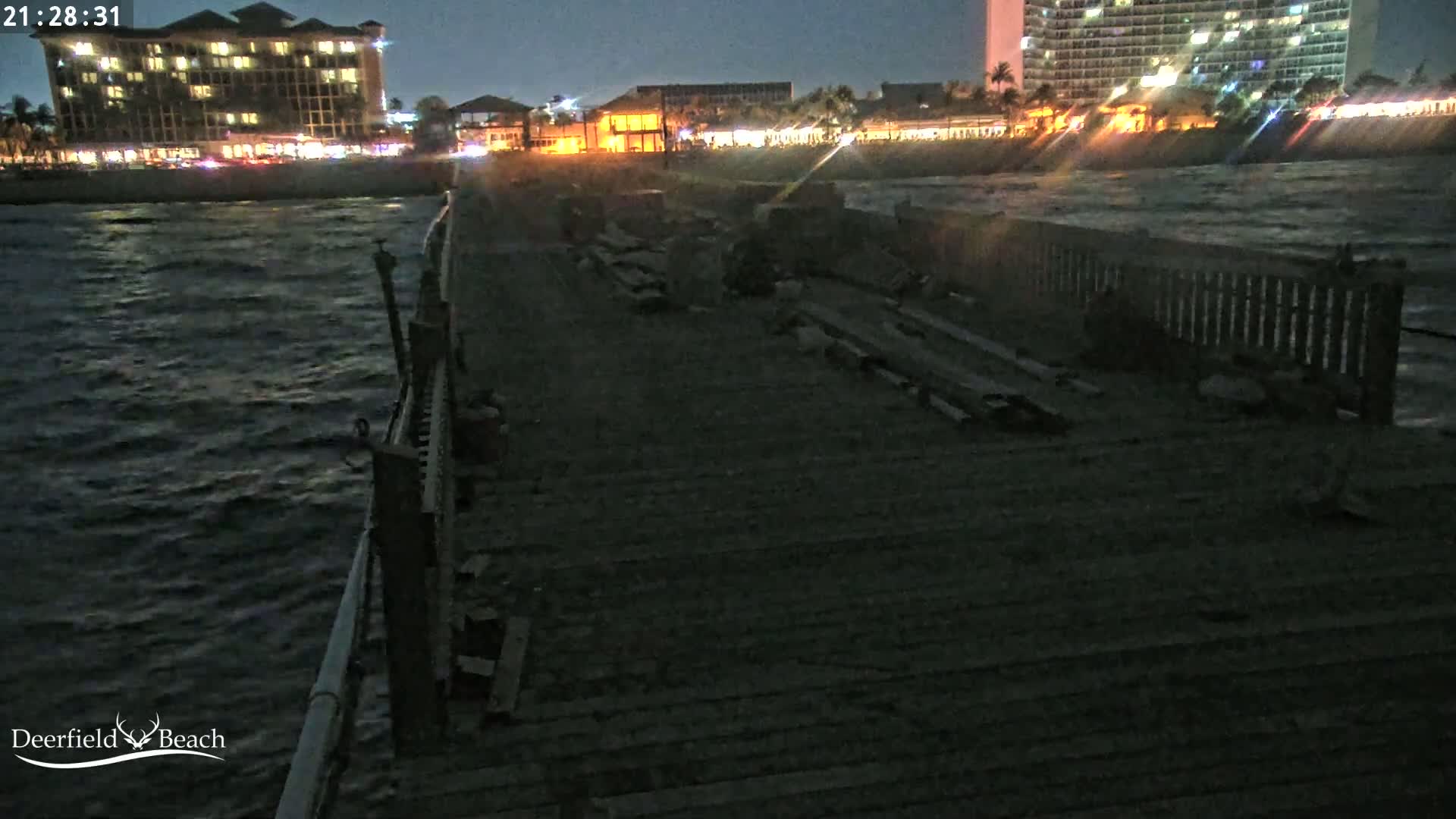 A dark, nighttime view of a partially damaged wooden pier extending over a choppy ocean, with illuminated buildings visible in the background.