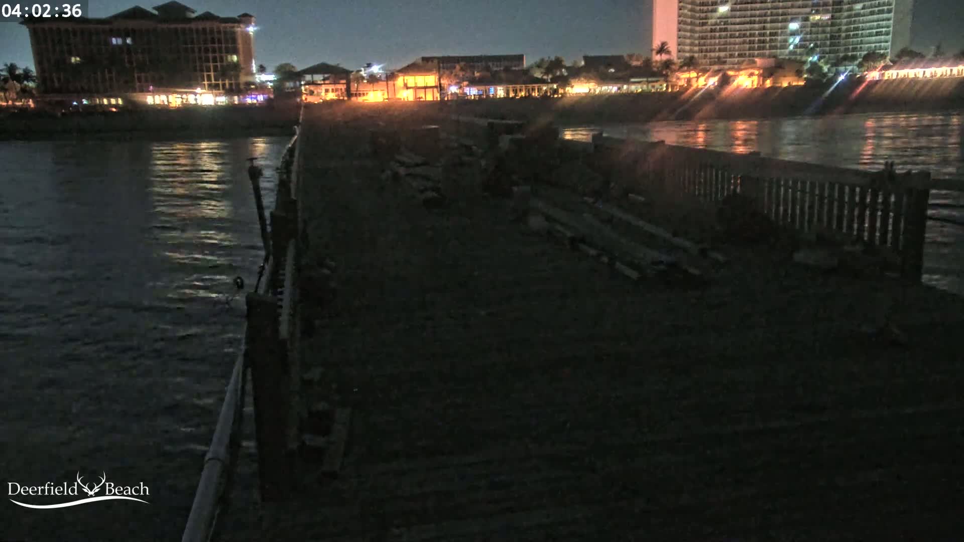 A dark, nighttime view from a pier shows calm water reflecting lights from buildings on the shore.