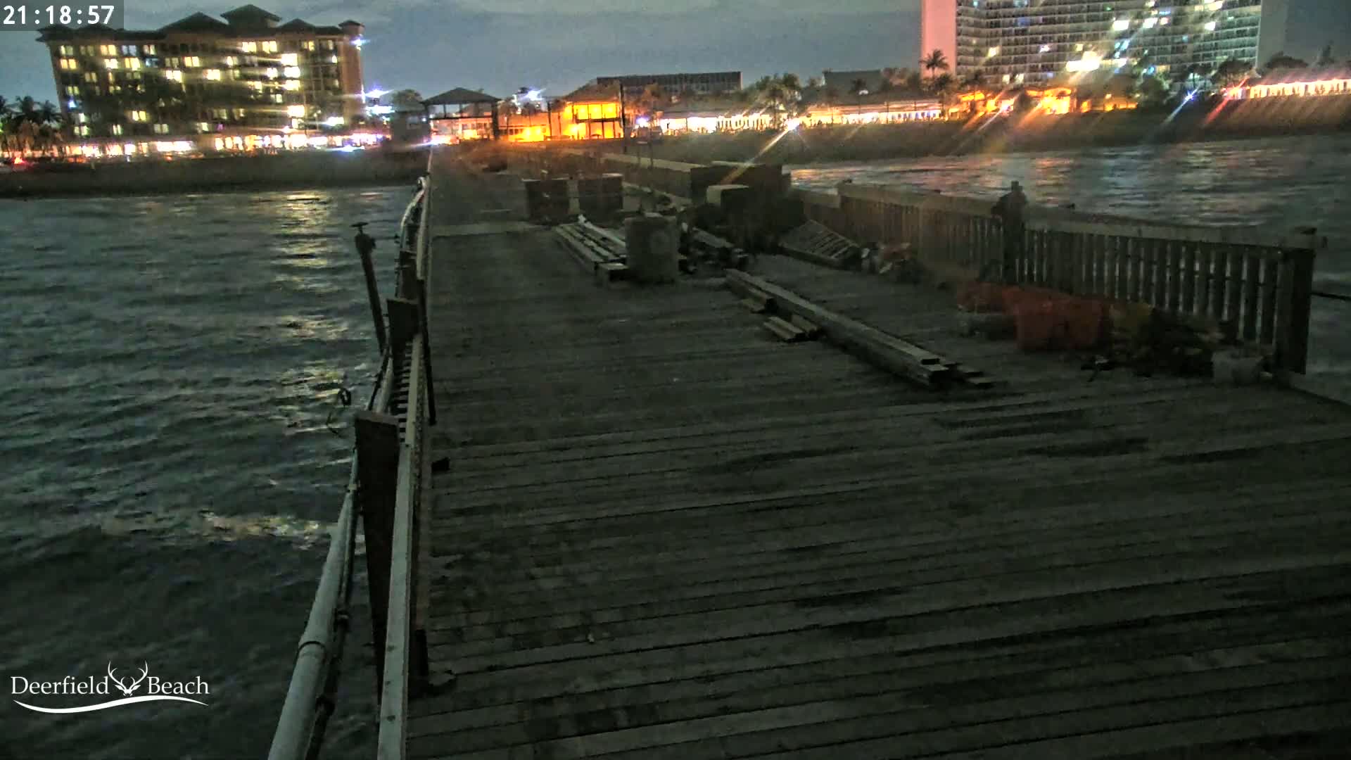 A dark wooden pier extends over a choppy, nighttime ocean toward a brightly lit shoreline with buildings.