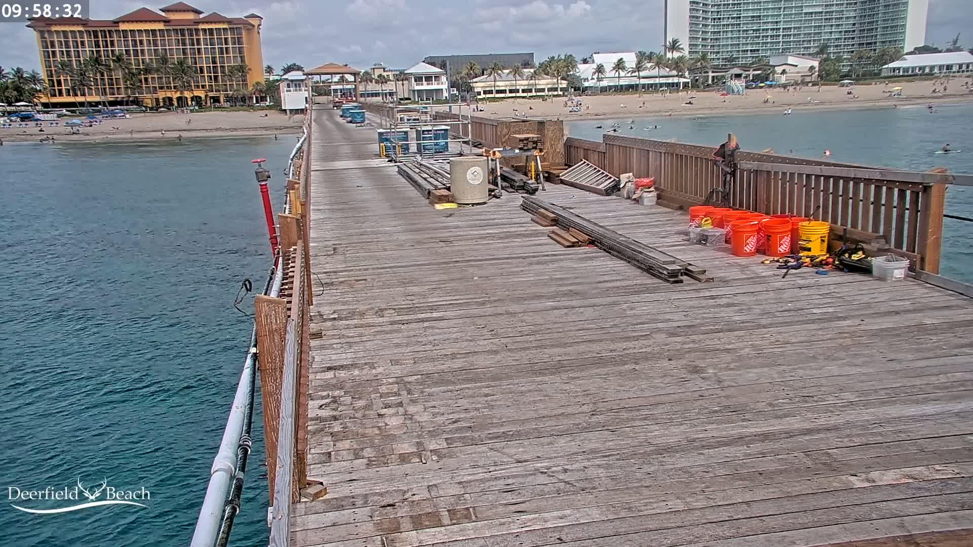A mostly sunny day shows a wooden pier under construction, with building materials and tools scattered on the deck, overlooking a calm ocean with a beach and hotels in the background.