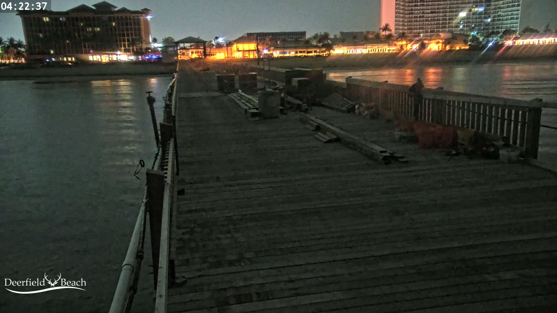 A dark wooden pier at night extends into a calm body of water, with construction materials scattered along it, and a brightly lit hotel and buildings visible in the background.