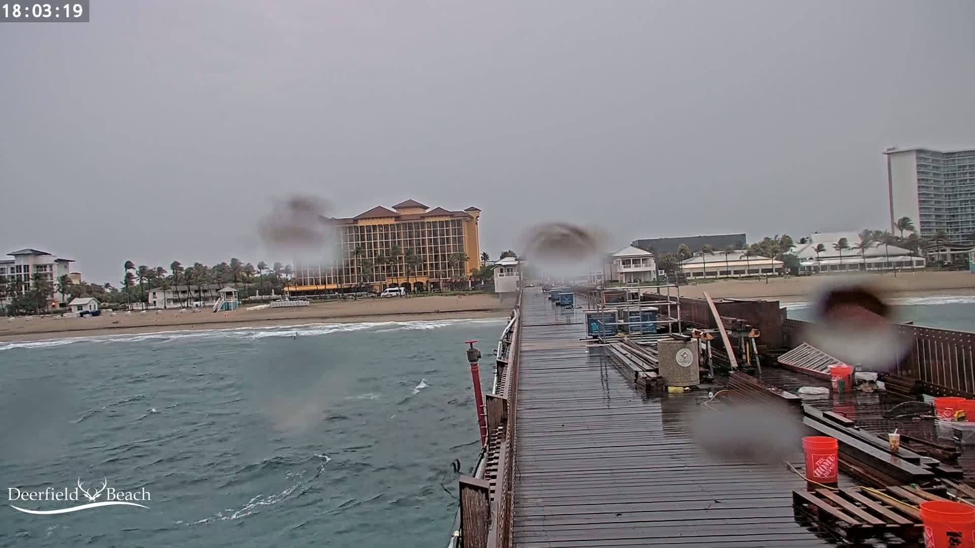 A rain-soaked pier extends from the frame toward a beach with hotels and buildings, under an overcast sky.