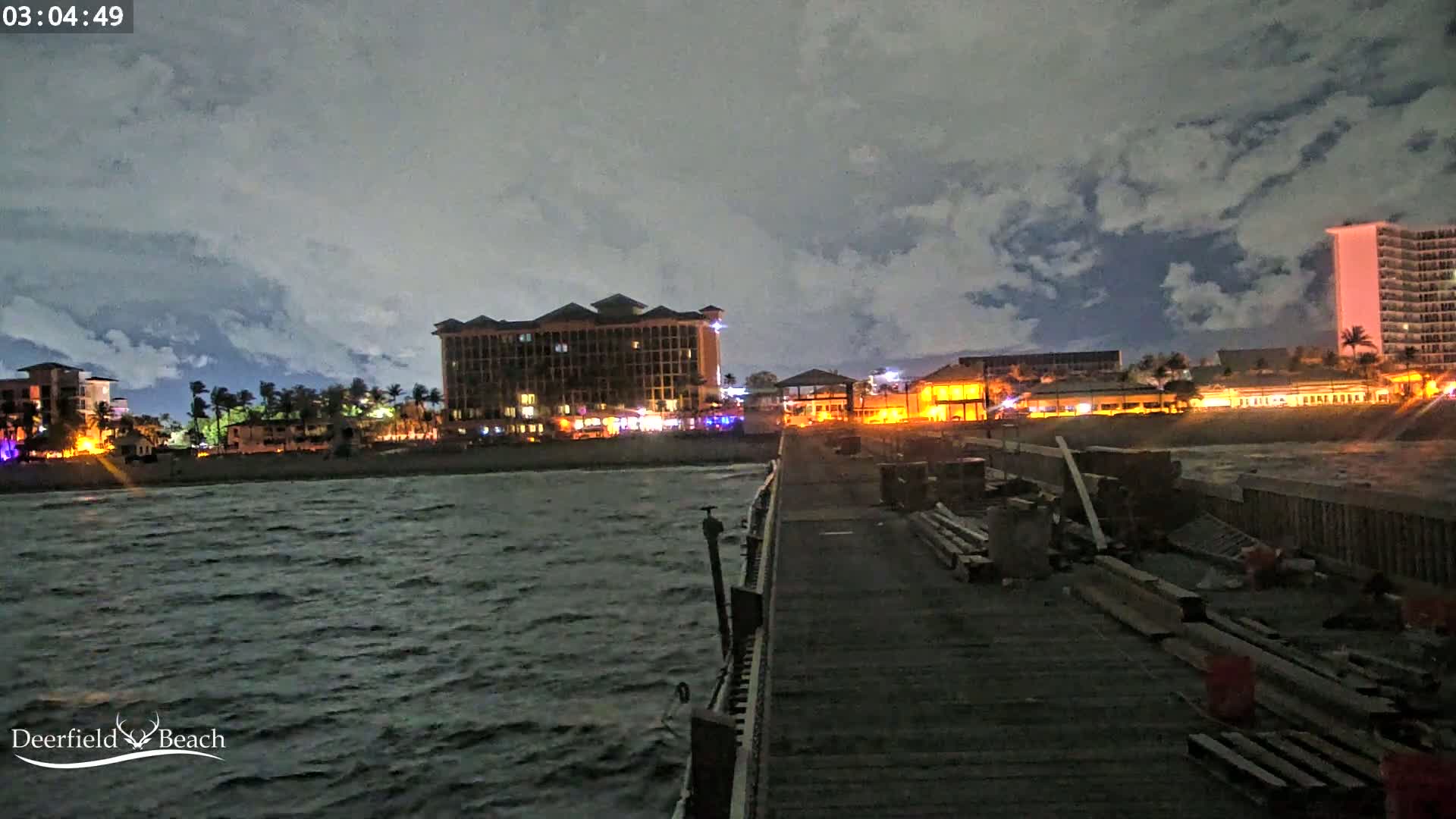 A nighttime view from a pier shows a beach with illuminated buildings under a cloudy sky and a choppy sea.