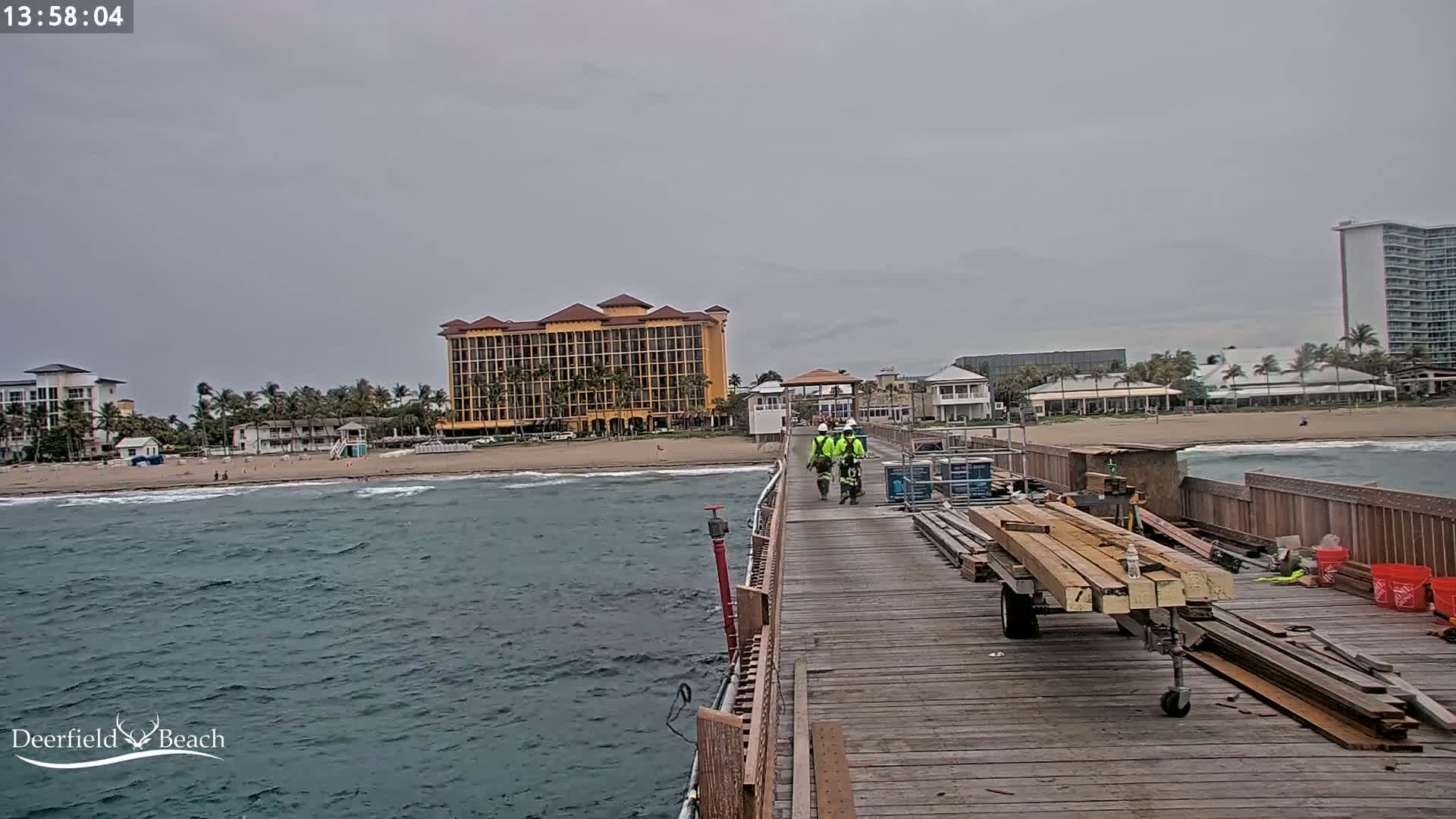 Three people walk down a pier toward a beach with moderate waves under an overcast sky, past construction materials and a large hotel.