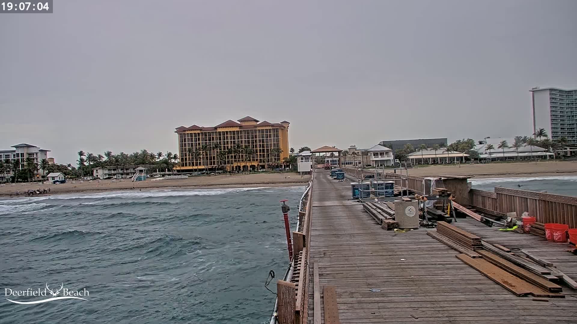 A wooden pier extends over a wavy ocean toward a beach with hotels and buildings under an overcast sky.