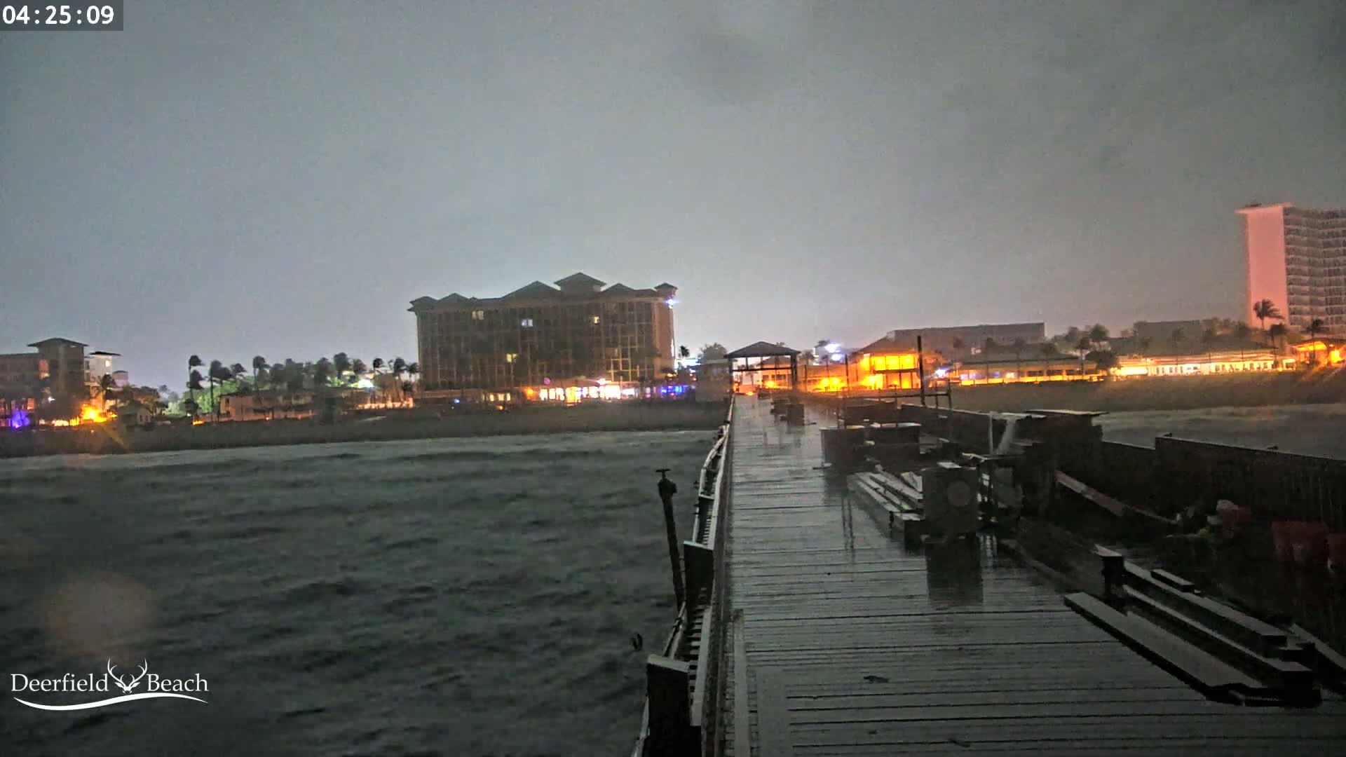 A rain-soaked pier extends into a dark, choppy ocean under a stormy night sky, with illuminated buildings visible in the background.