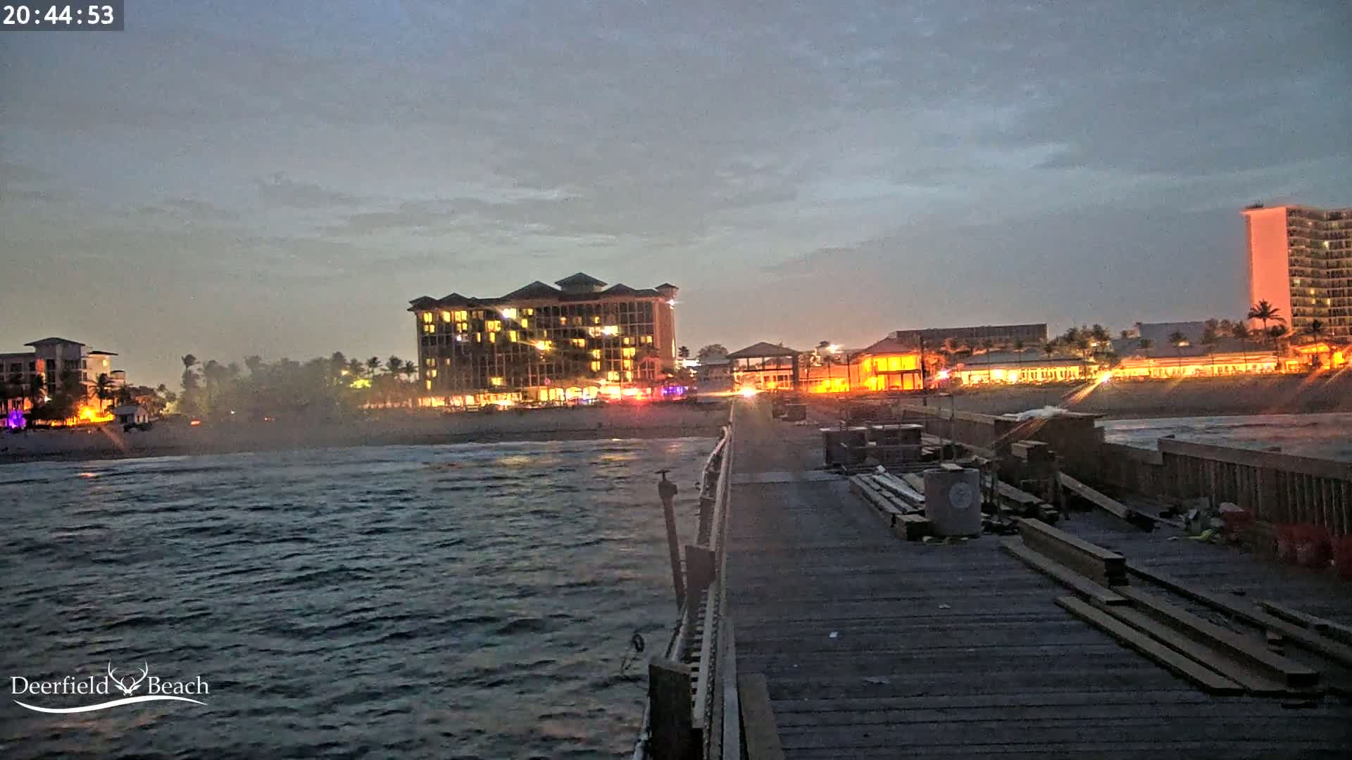 A view from a pier at night shows a dark ocean, a beach with illuminated buildings, and construction materials on the pier under a twilight sky.