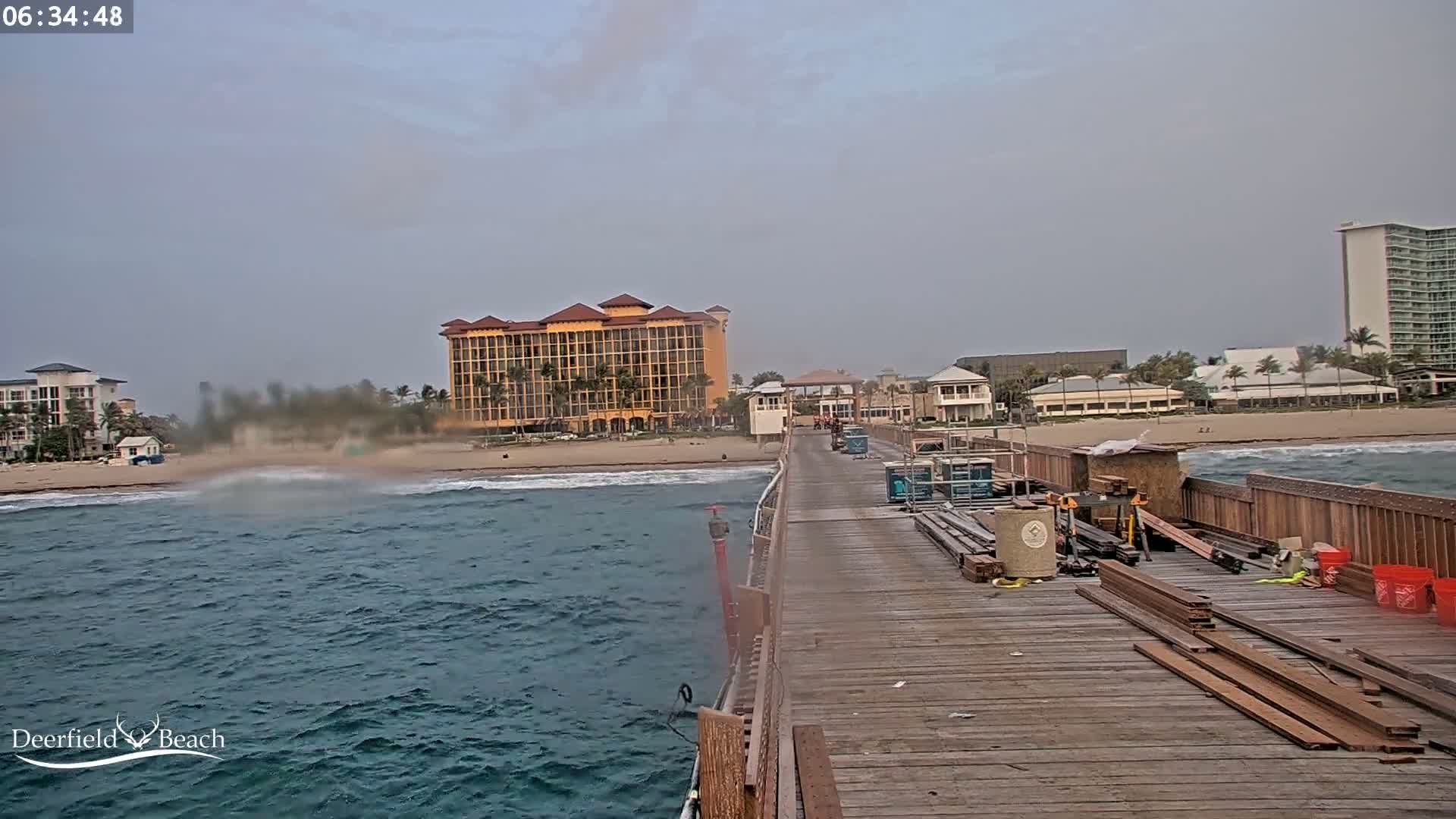 A wooden pier under a hazy sky extends over a choppy ocean toward a beach with hotels and buildings.