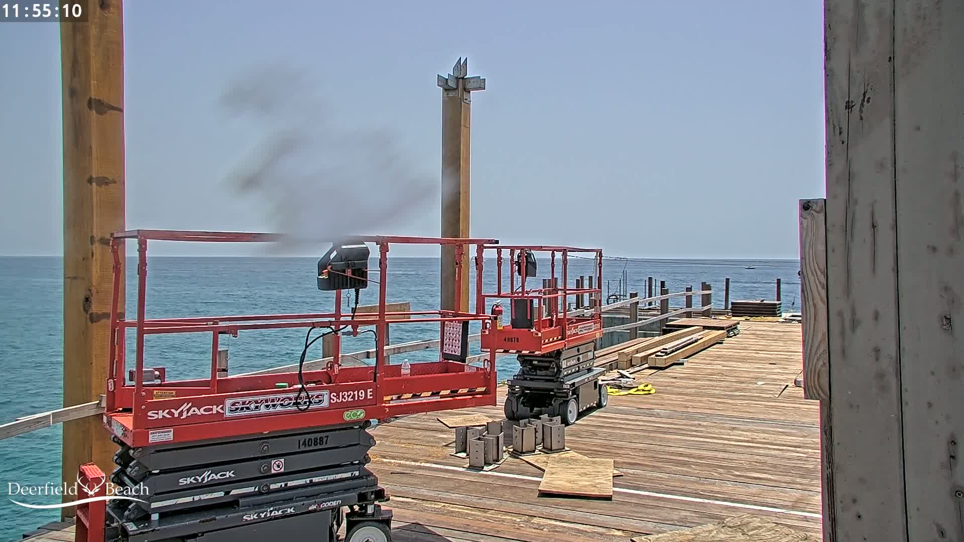 Two red scissor lifts are on a wooden pier overlooking a calm ocean under a clear sky, with construction materials and a wooden post nearby.