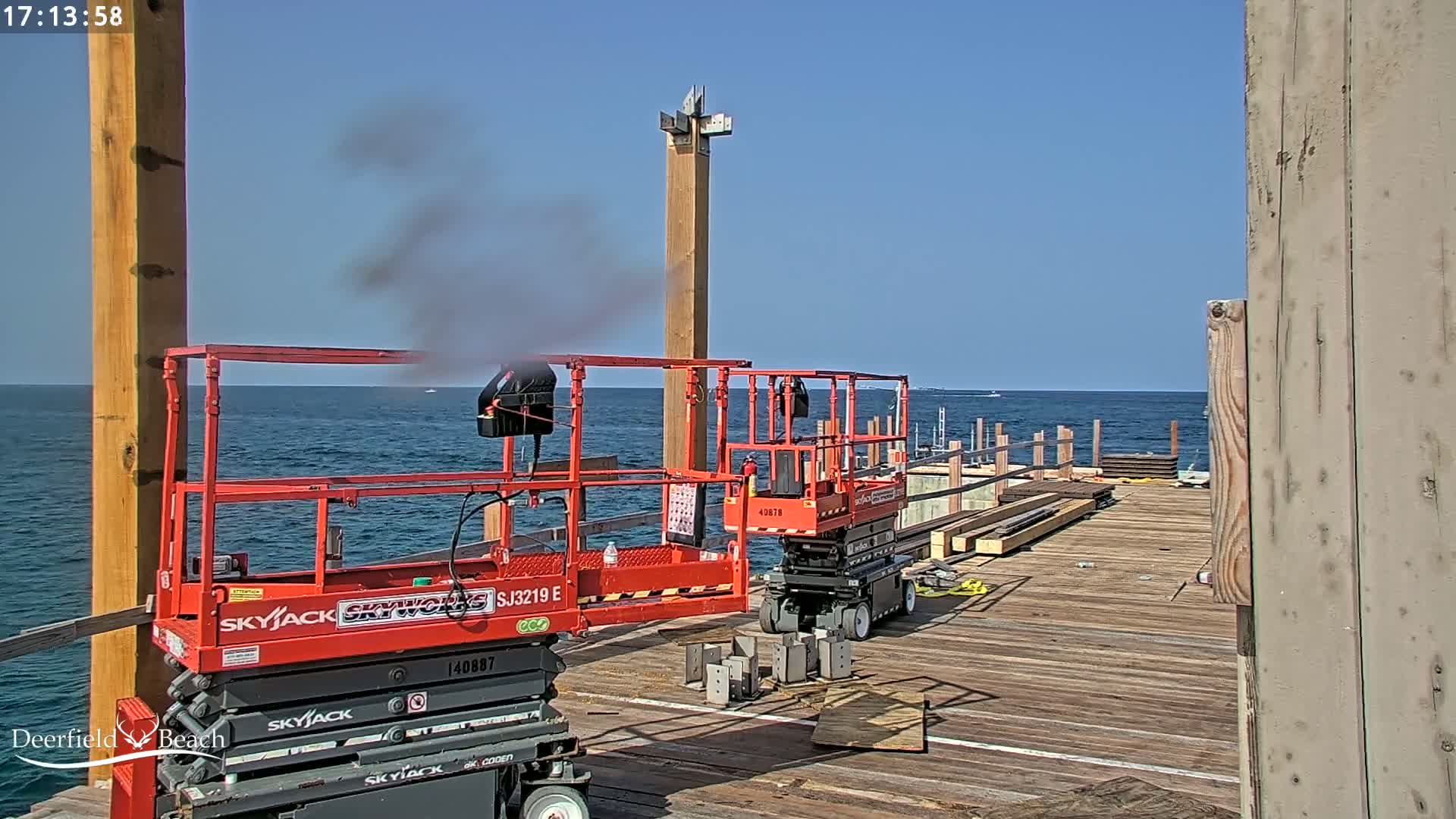 Two red aerial work platforms are positioned on a wooden pier overlooking a calm ocean under a clear blue sky.