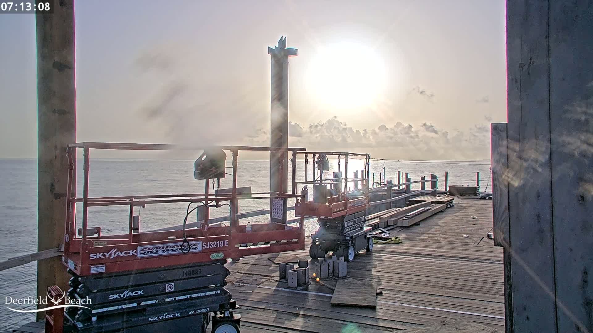 Construction equipment sits on a pier overlooking a calm ocean under a partly cloudy, sunrise sky.