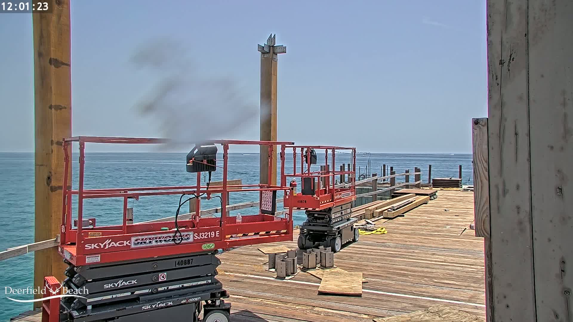 Two red scissor lifts are on a pier under construction over a calm ocean on a sunny day.