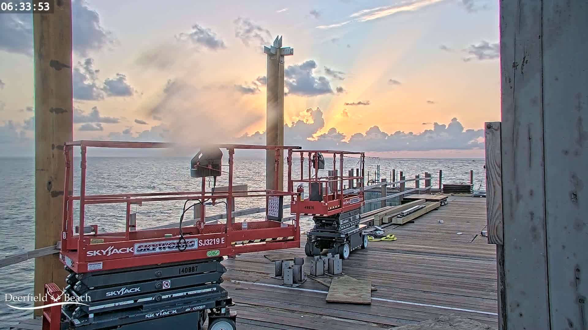 Two red Skyjack scissor lifts are parked on a wooden pier during a sunrise over a calm ocean.