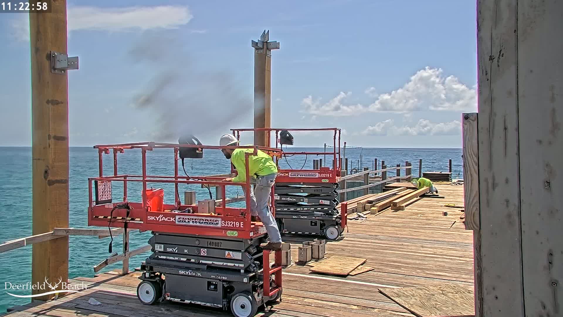 Two workers on scissor lifts are constructing a pier on a sunny day with partly cloudy skies over a calm ocean.