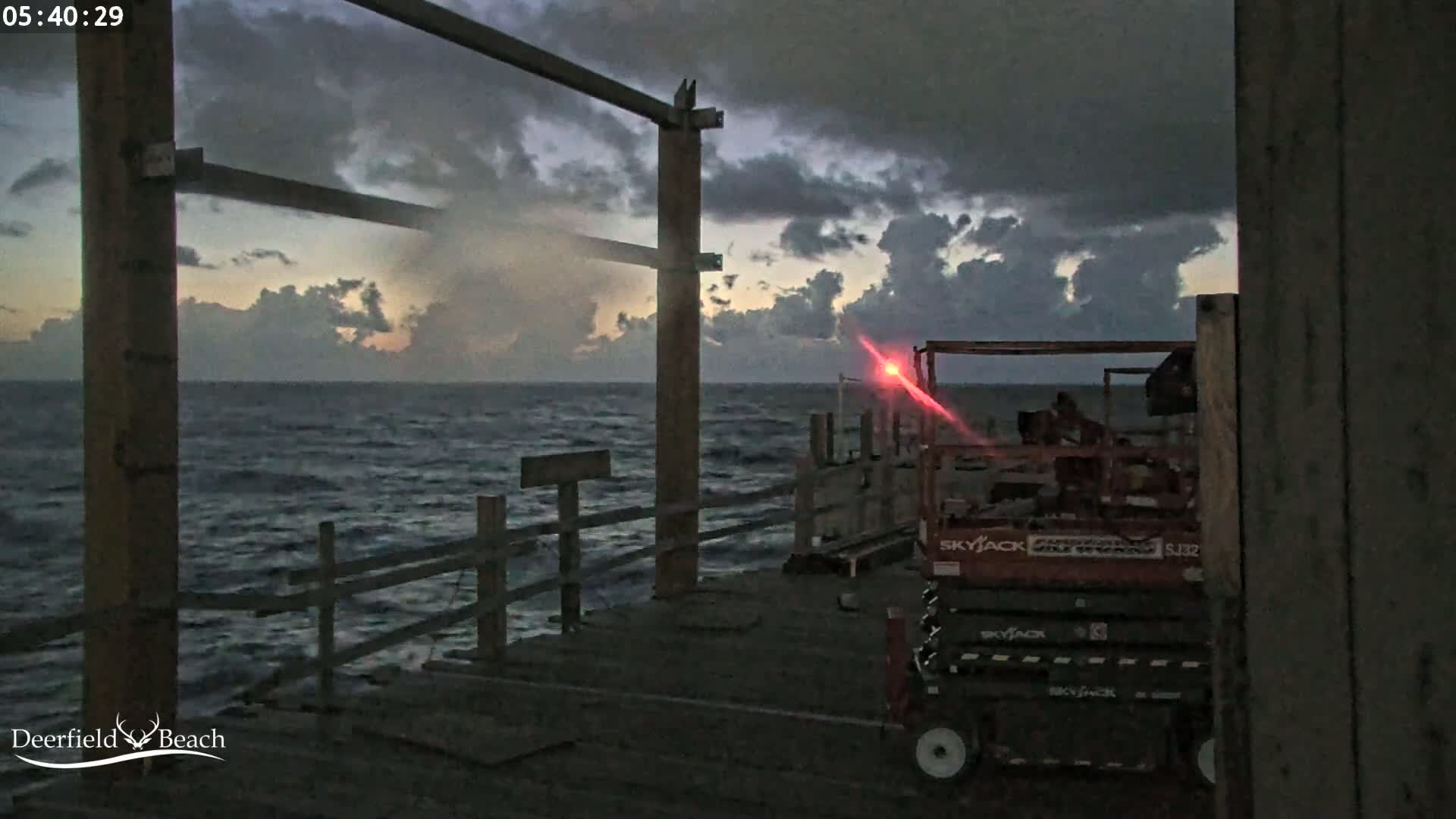 A partially constructed pier overlooks a dark, choppy ocean under a cloudy, pre-dawn sky, with a red light visible in the distance and a piece of construction equipment on the pier.