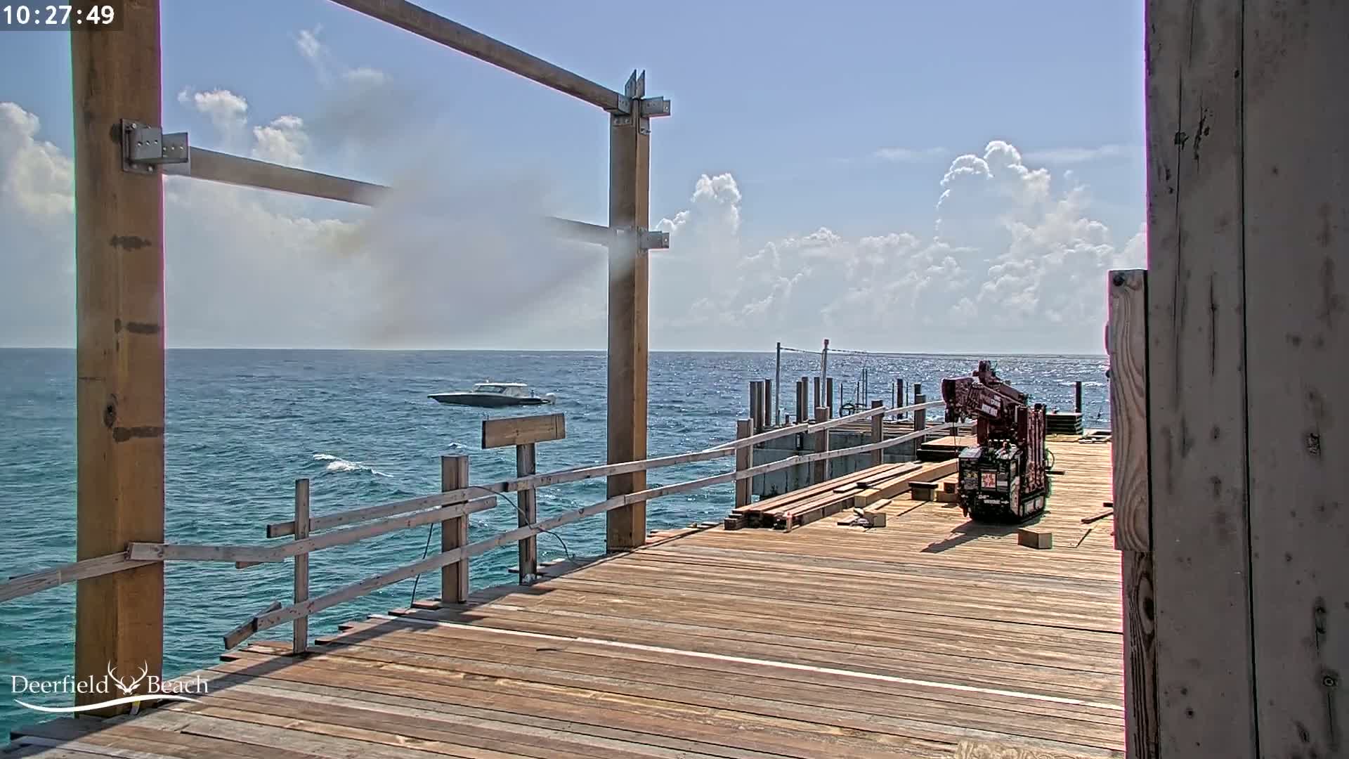 A partly cloudy day shows a wooden pier under construction, with a small boat on the ocean in the background and a crane on the pier.