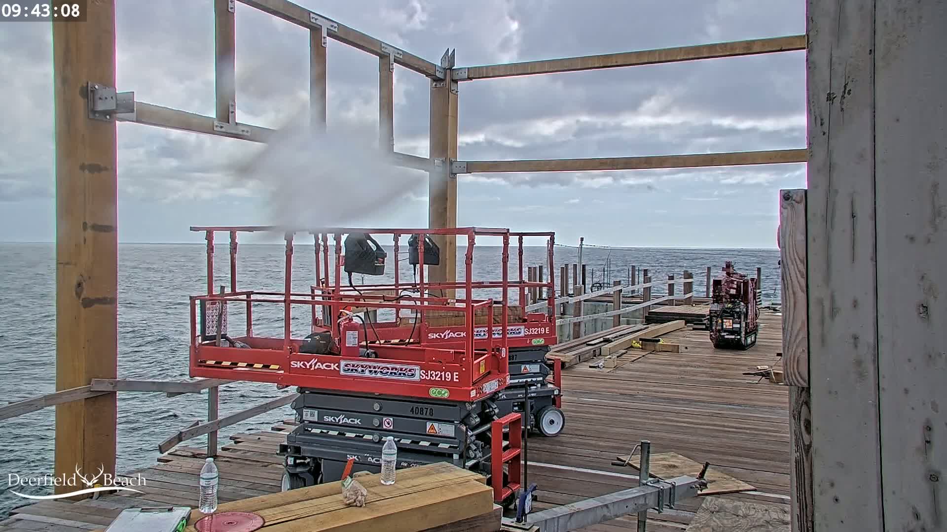 Two red Skyjack scissor lifts sit on a wooden pier overlooking a calm ocean under an overcast sky, with one emitting steam.