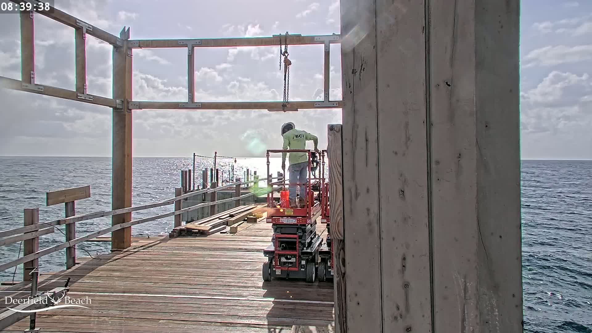A construction worker on a lift operates machinery on a wooden pier overlooking a calm ocean under a partly cloudy sky.