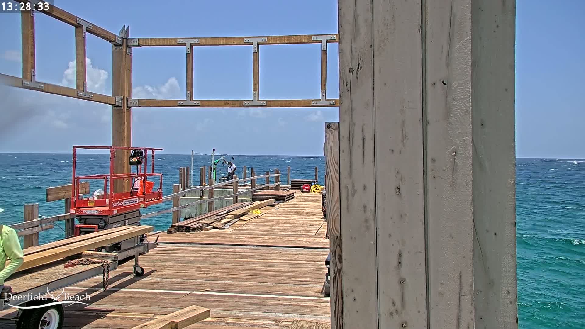 A partially constructed wooden pier extends over a blue ocean with small waves under a partly sunny sky, featuring construction equipment and workers.