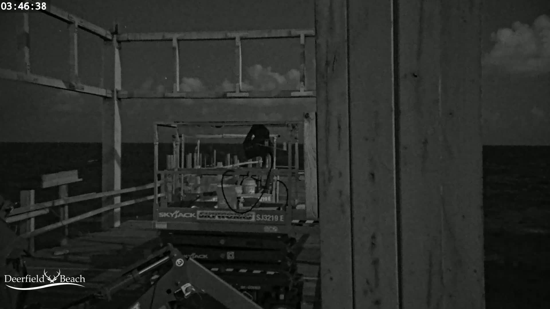 A nighttime, black-and-white image shows a section of a pier under construction, with a small piece of construction equipment visible, and a dark body of water in the background under a partly cloudy sky.